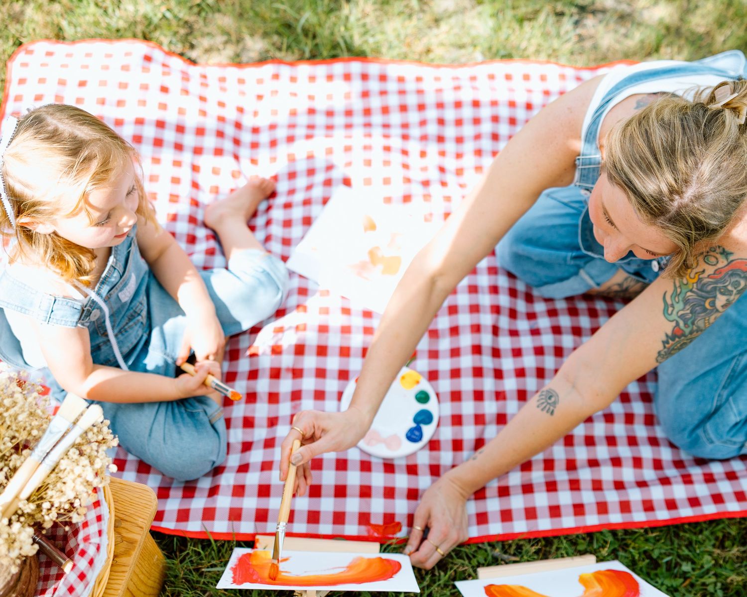 Mother and daughter painting together on a red checkered picnic blanket outdoors.