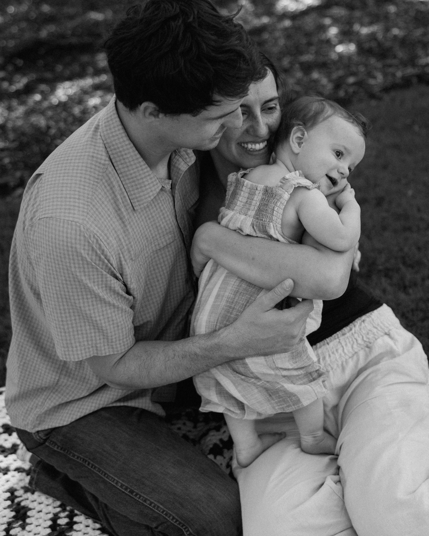 Black and white photo of a happy couple sitting outdoors, smiling and holding their laughing baby together.