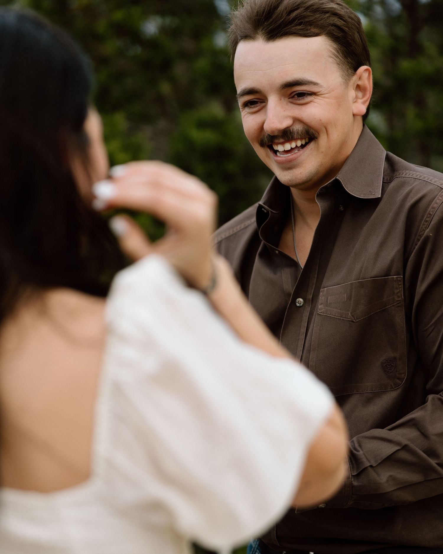 Smiling man with mustache in brown shirt laughing during outdoor couples engagement photo session.