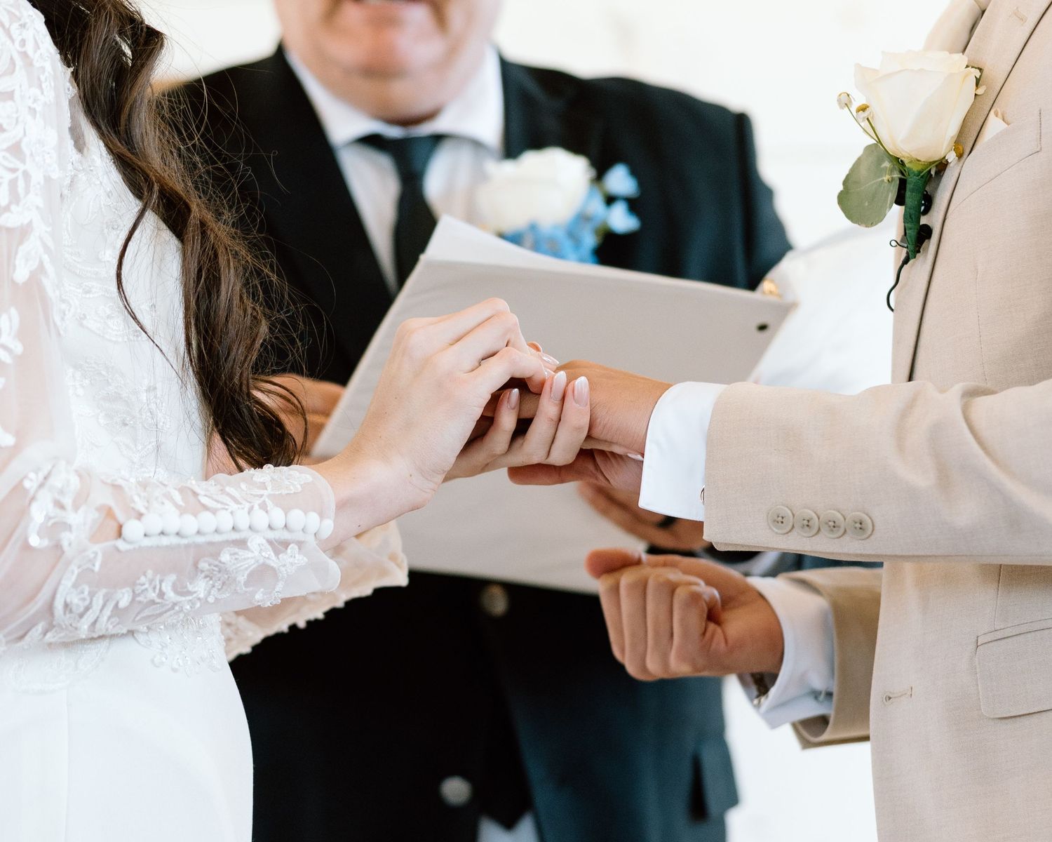 Bride placing wedding ring on groom's finger during ceremony, officiant holding book in background.