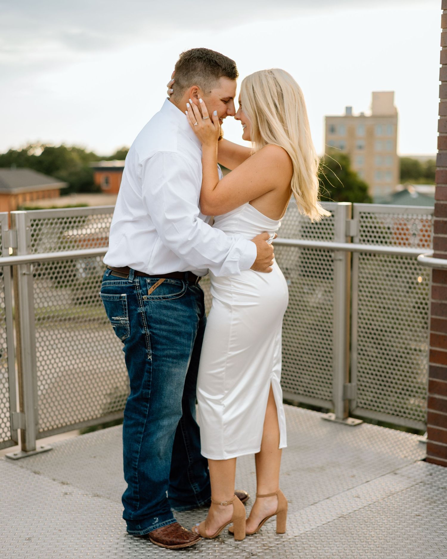 Couple in white outfits sharing a romantic moment on an outdoor rooftop bridge with urban buildings in the background.