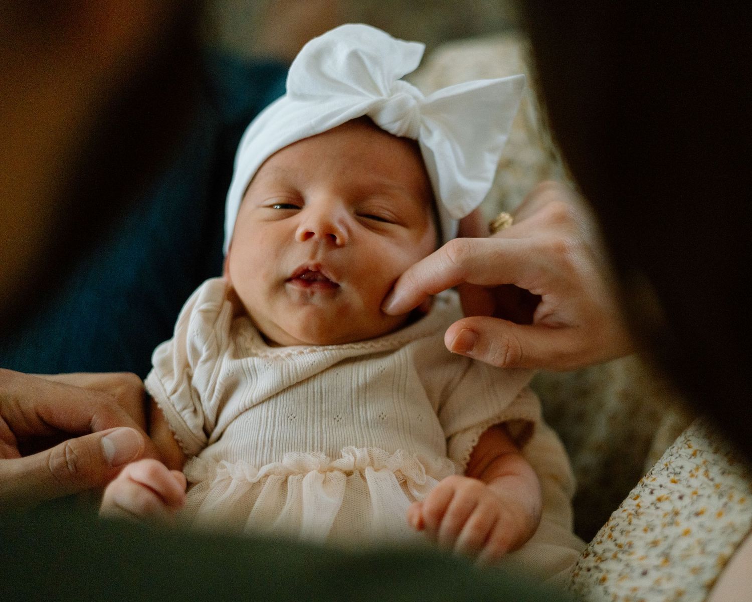 A sleeping newborn baby wearing a white bow and cream dress lies peacefully while being touched by gentle hands.