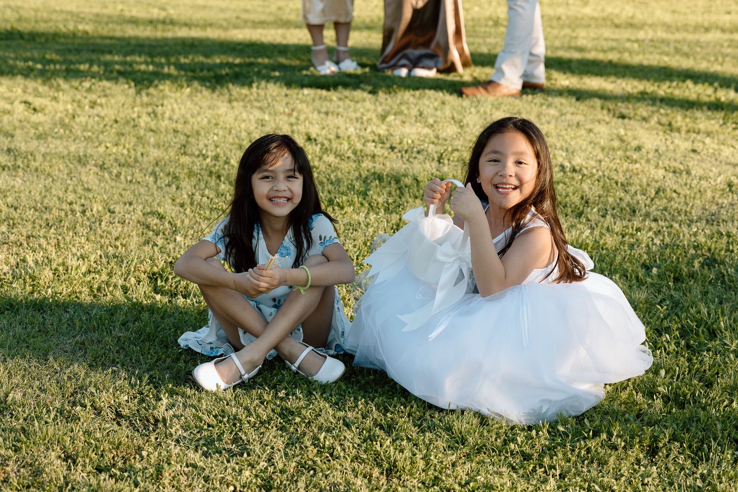 Young wedding attendants sit together on grass during outdoor reception celebration.