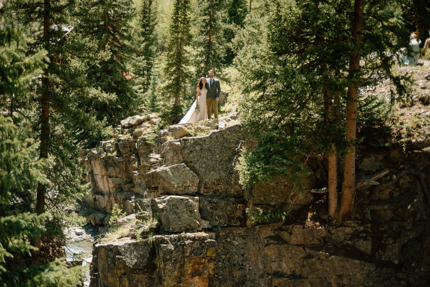 A couple stands atop a dramatic rocky cliff surrounded by evergreen trees in a mountain landscape.