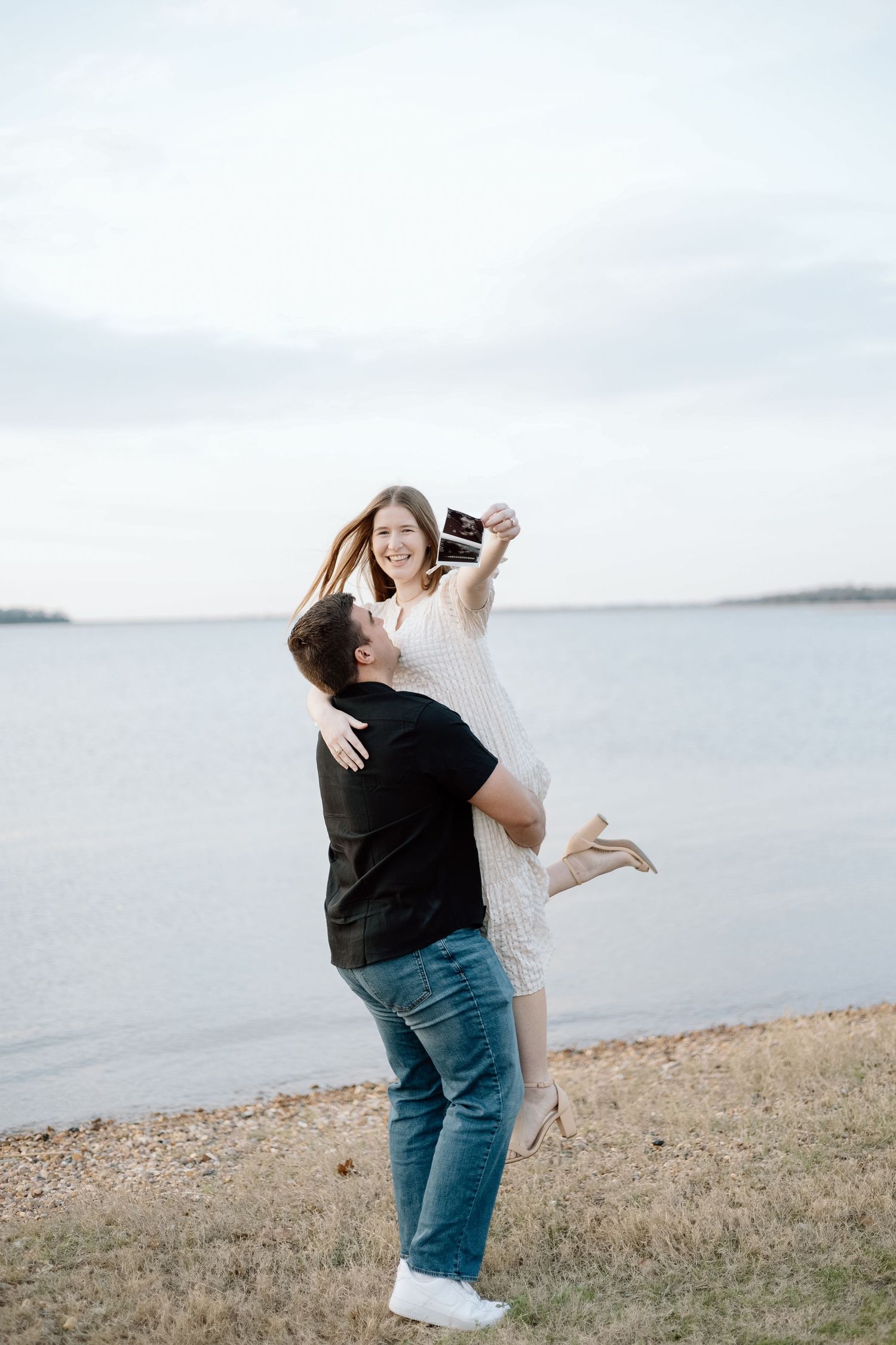 Couple celebrates pregnancy announcement with joyful lift pose by waterfront.
