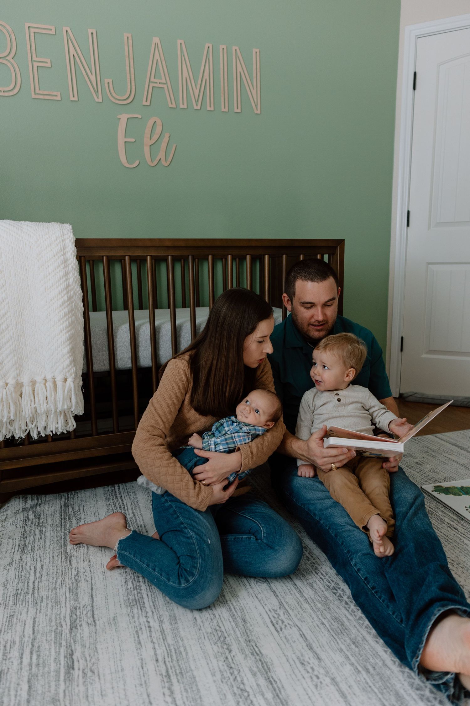 A family sits together on the floor of a nursery reading a book, with the name Benjamin displayed on the mint green wall.