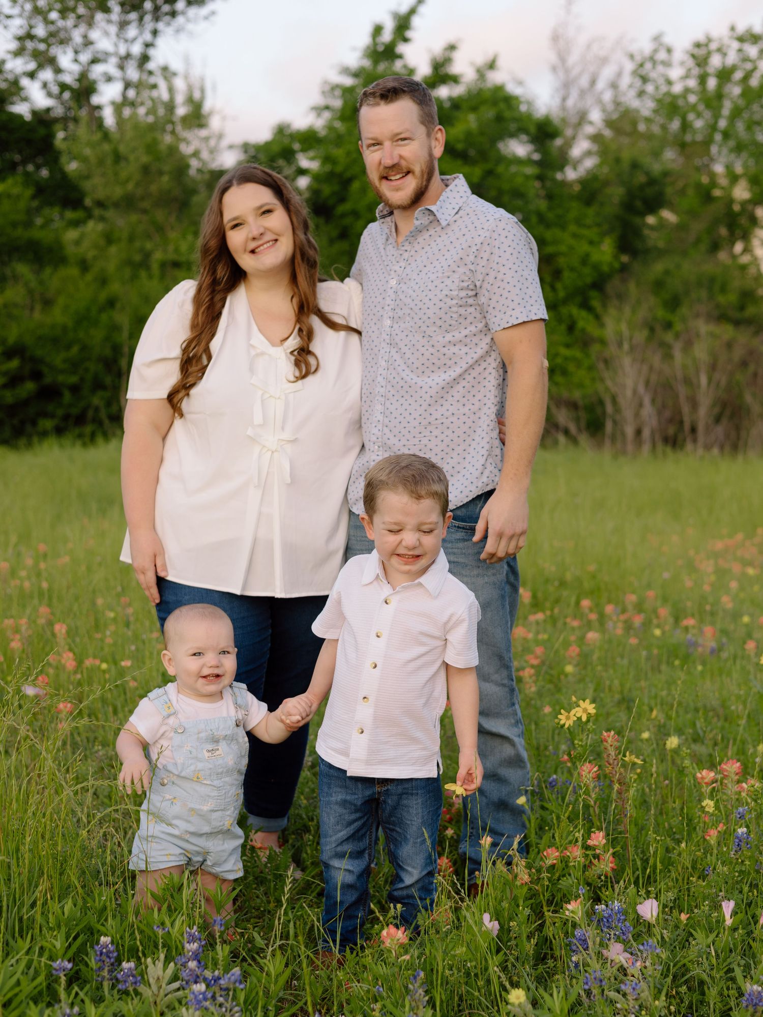 A family poses together in a wildflower meadow during springtime, wearing coordinated white and casual outfits.