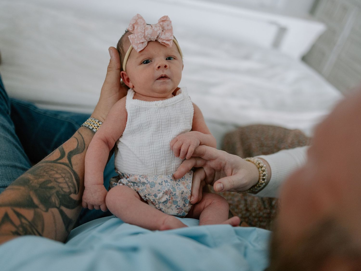 A baby wearing a white sleeveless outfit and pink bow headband sits on a couch with tattooed hands nearby.