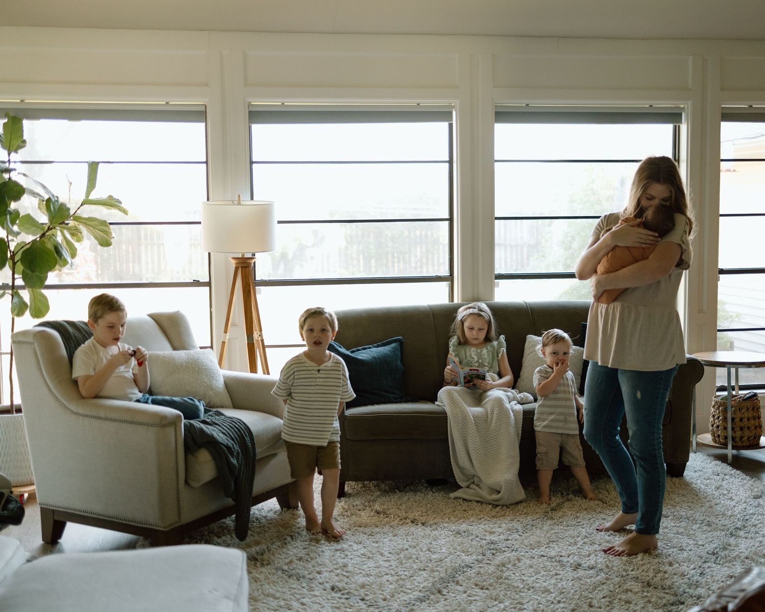 Family relaxes together in a sunlit living room with large windows and cozy furniture on a bright afternoon.