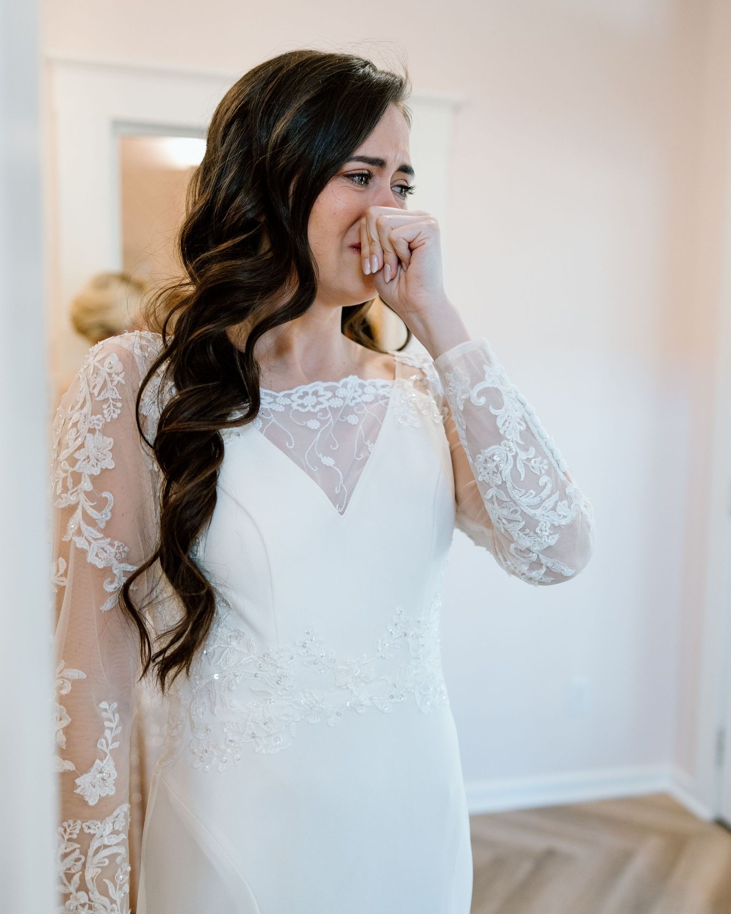 Emotional bride in lace long-sleeve wedding gown covering her mouth while getting ready.