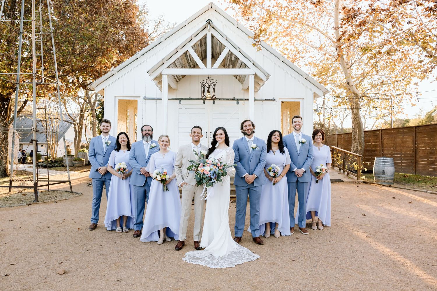Wedding party in blue dresses and tan suits posing before white barn chapel.