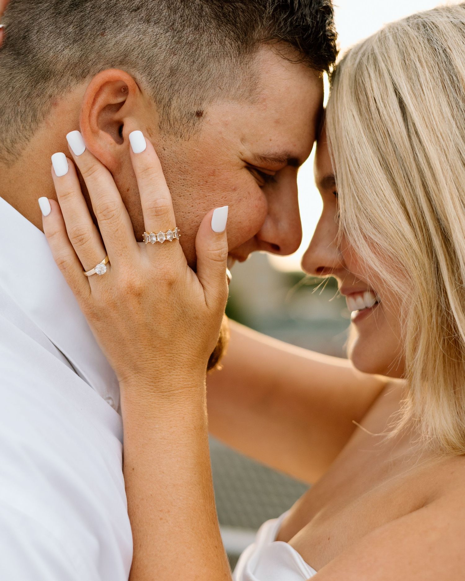 Close-up of smiling couple touching foreheads, woman displaying elegant diamond engagement ring with white manicured nails.
