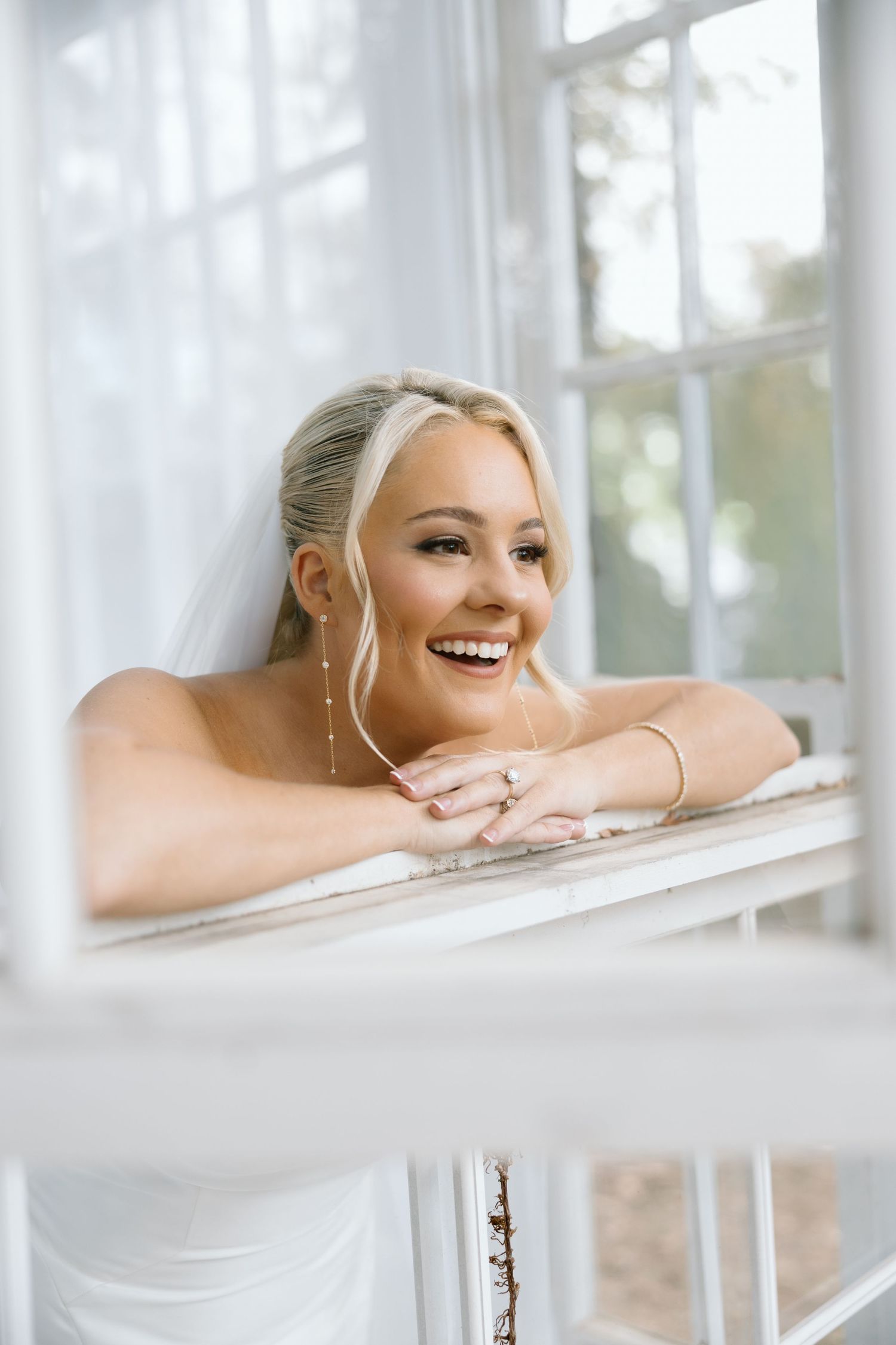 Smiling blonde bride in veil leans on white railing by window, wearing pearl earrings and bracelet on wedding day.