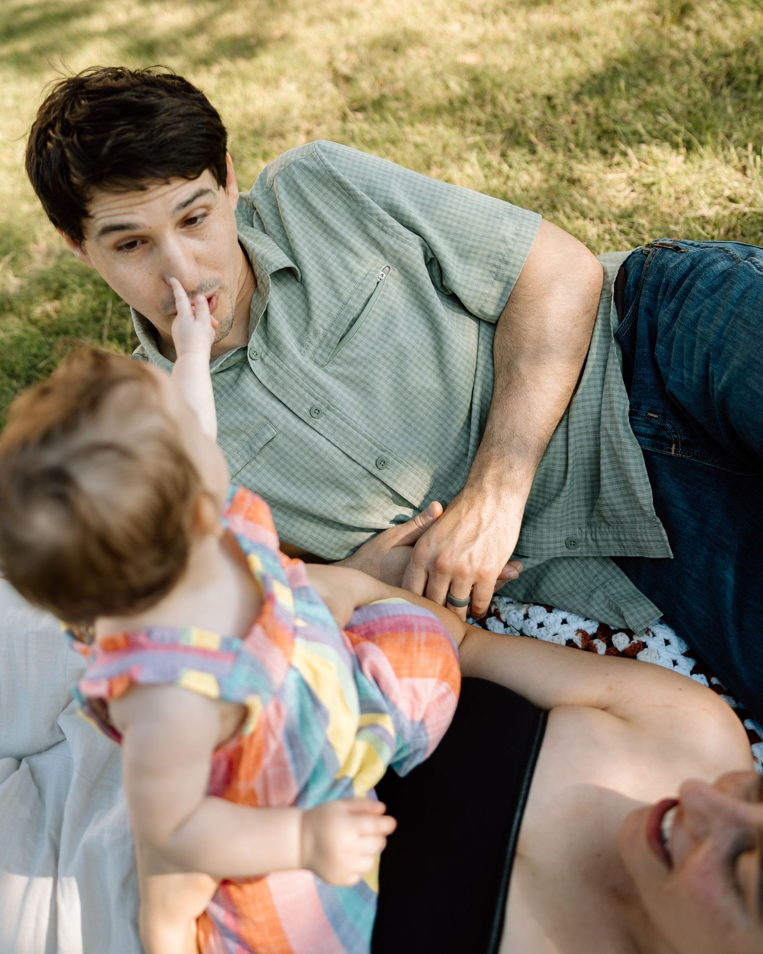 A man lying on grass interacts with a toddler in a colorful outfit while another person laughs nearby.