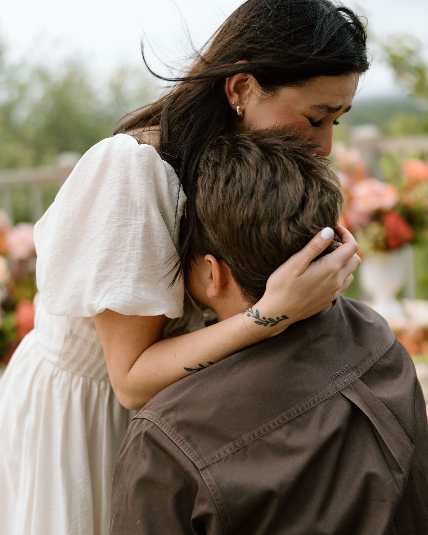 A woman in a white dress tenderly embraces a person in a brown jacket outdoors near blooming flowers.