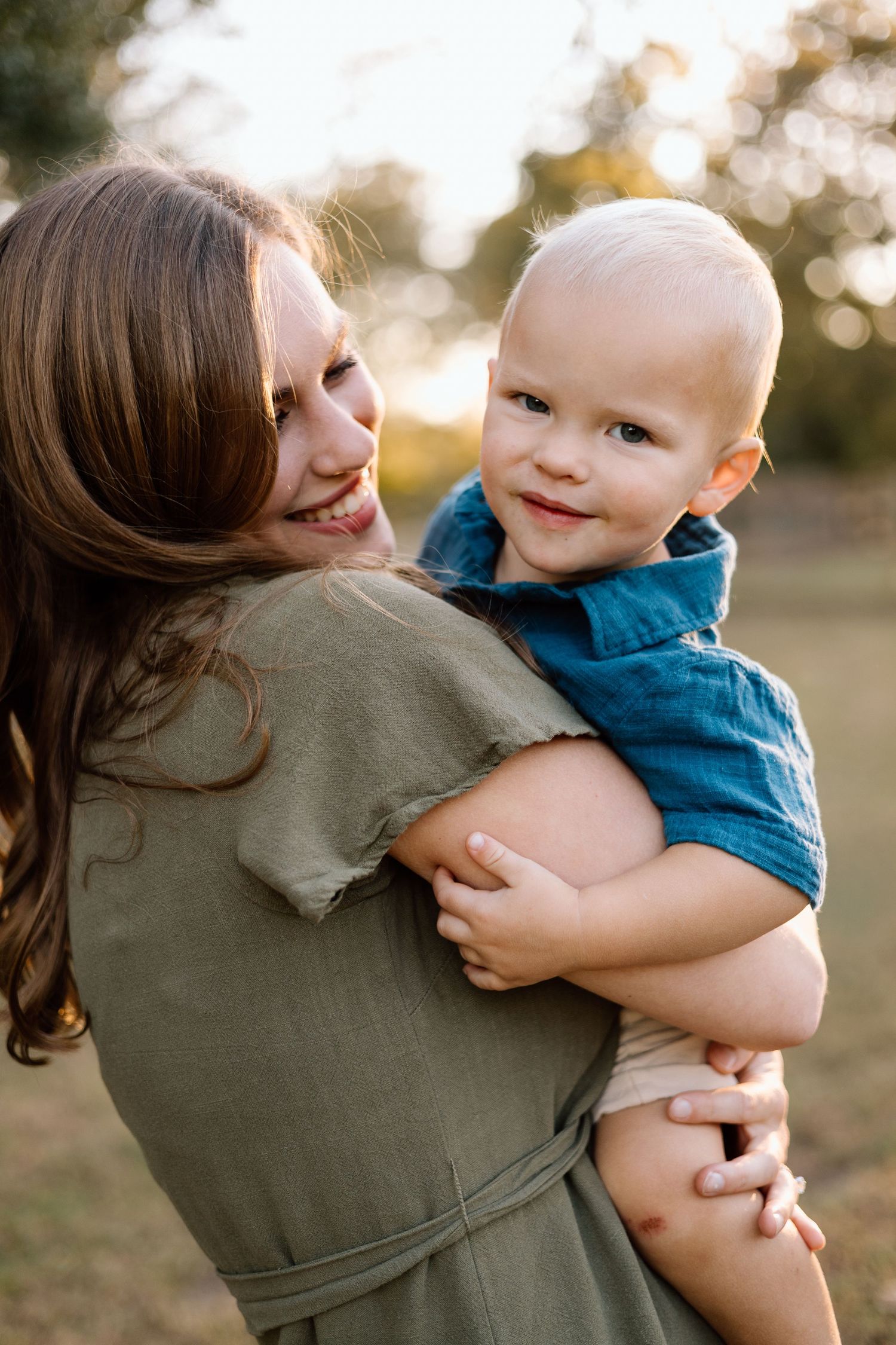 A smiling mother in olive dress holds her happy toddler boy outdoors in golden hour sunlight.