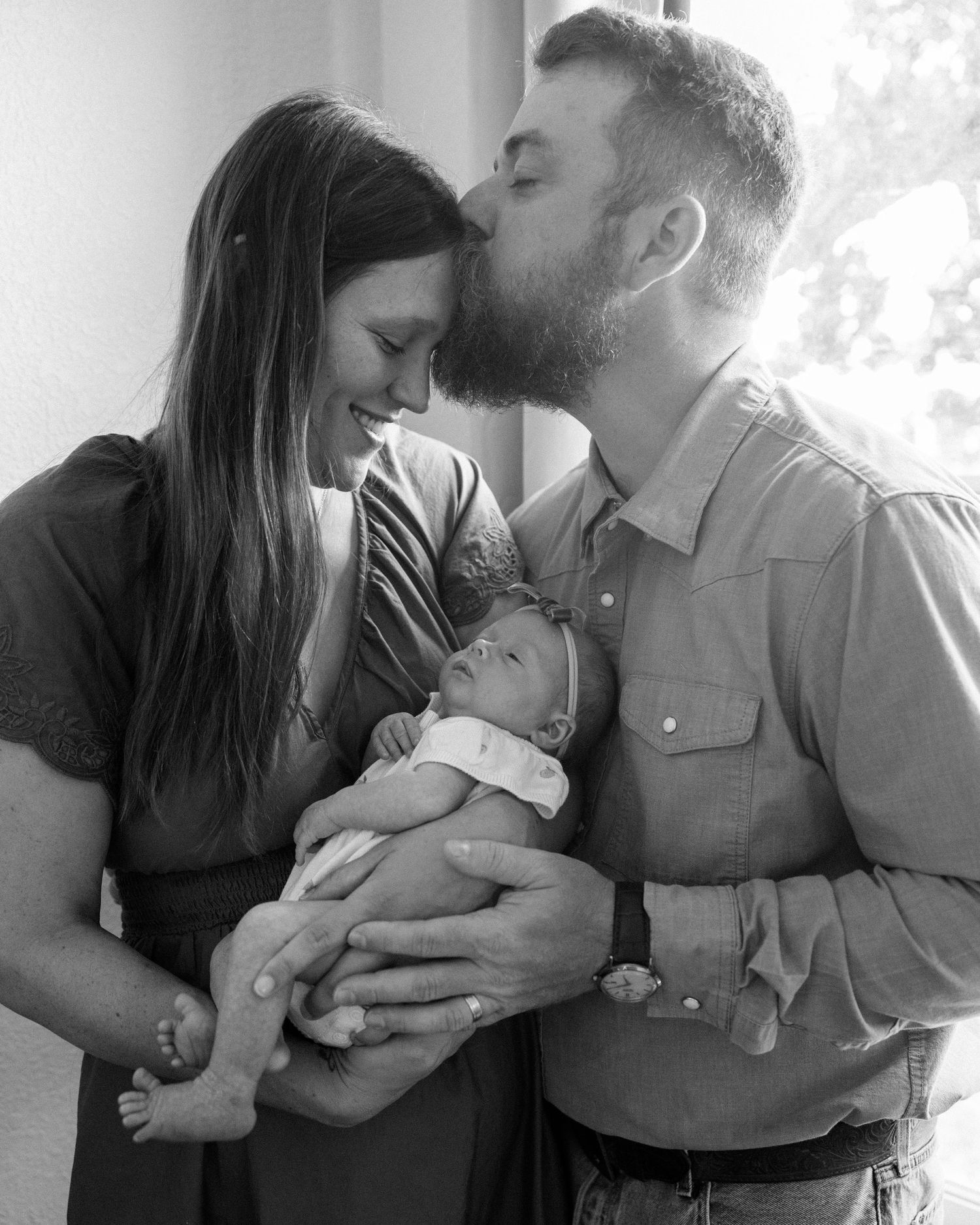 Black and white photo of a couple sharing a tender moment, kissing over their newborn baby held in their arms.