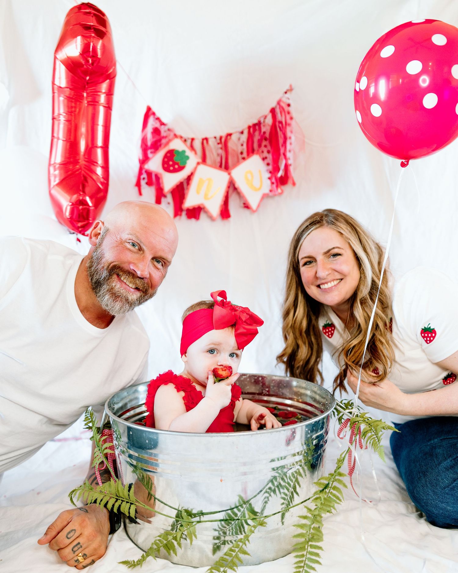 Family celebrating baby's first birthday with red balloons, strawberry banner, and infant in galvanized tub.