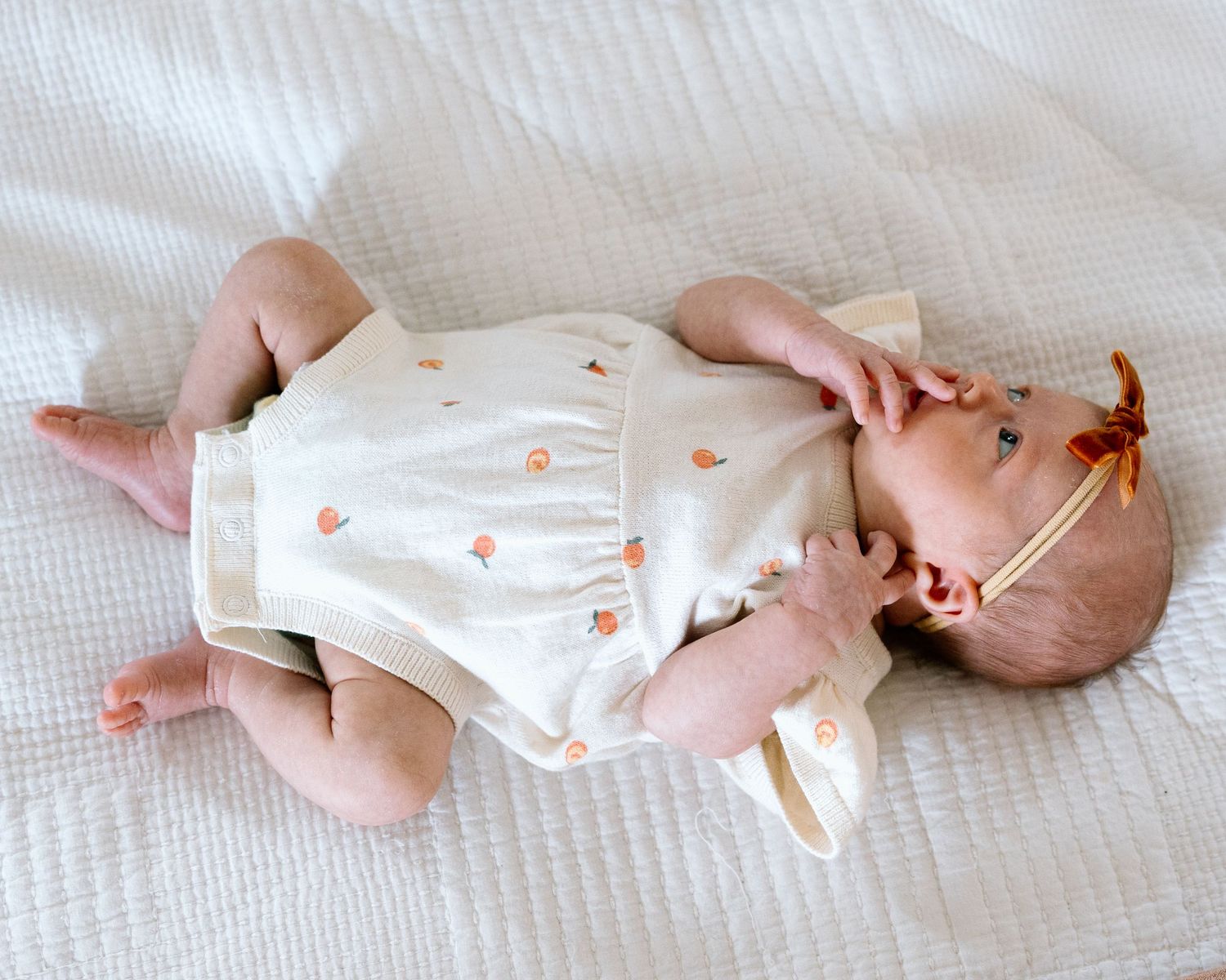 Baby girl in cream floral romper lying on white bedding, wearing a bow headband.