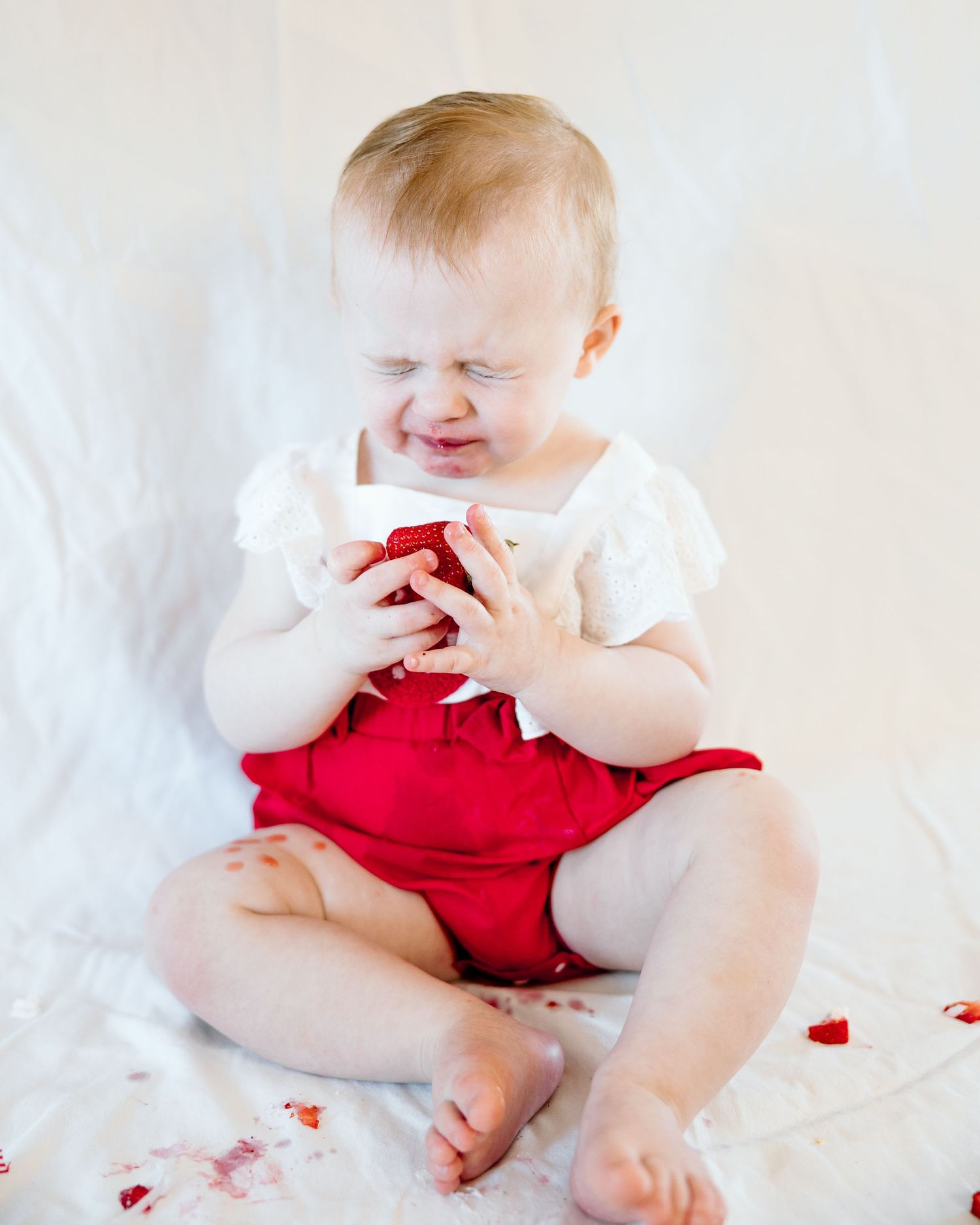 Cute baby in red romper and white top sitting on white blanket, smiling and holding a red object.