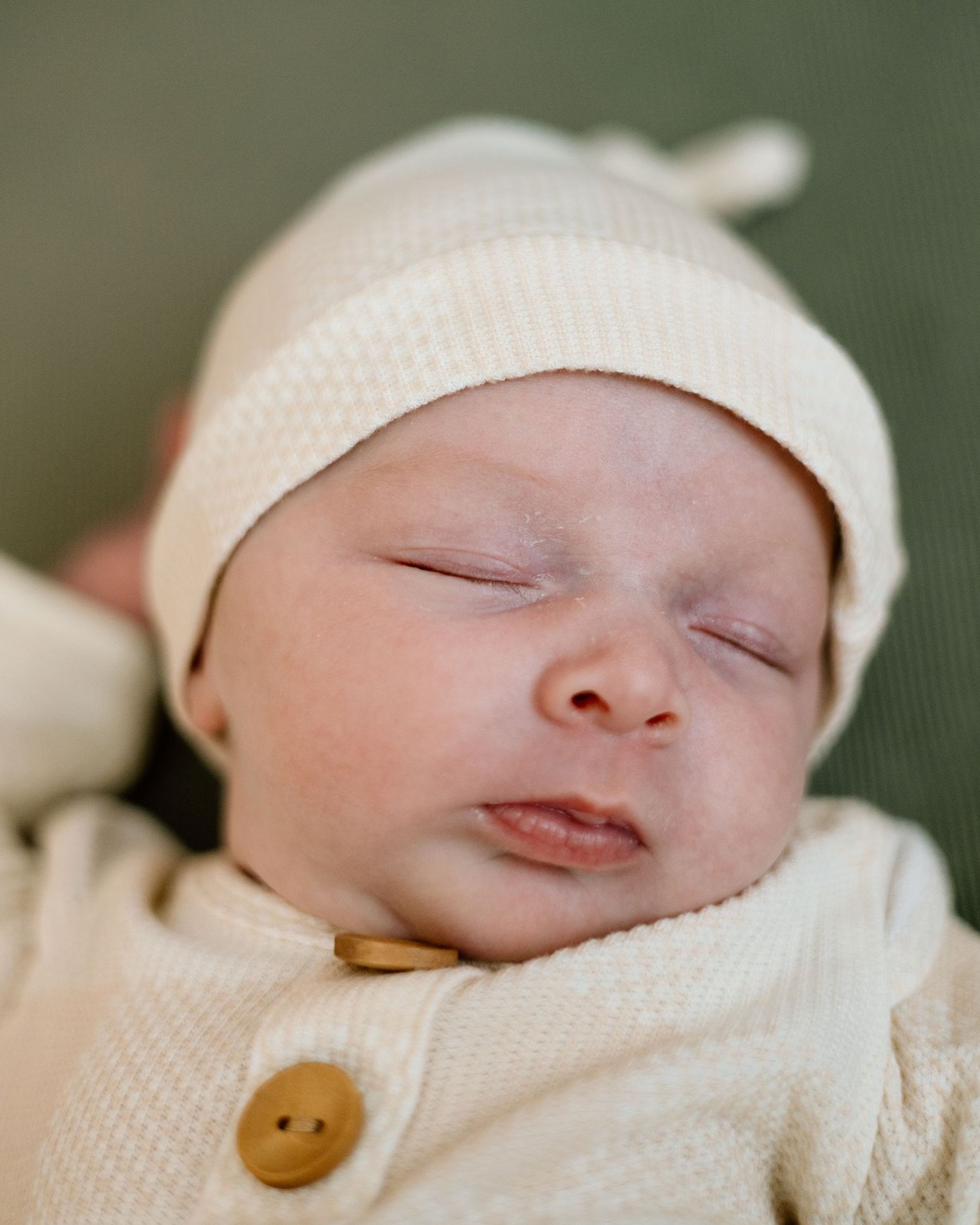 Peaceful sleeping newborn baby wearing cream knit hat and button onesie with eyes gently closed.