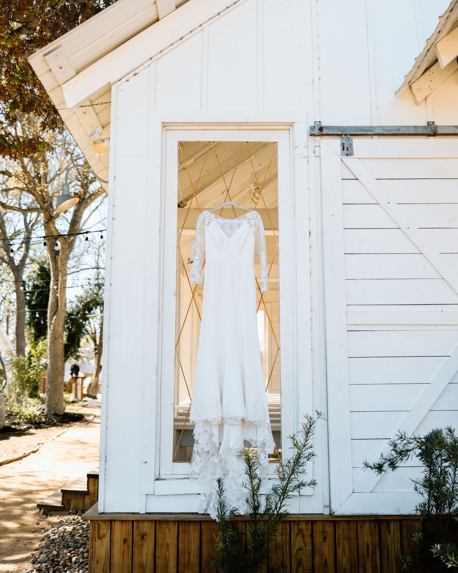 A white lace wedding dress hanging in a rustic white barn door frame outdoors.