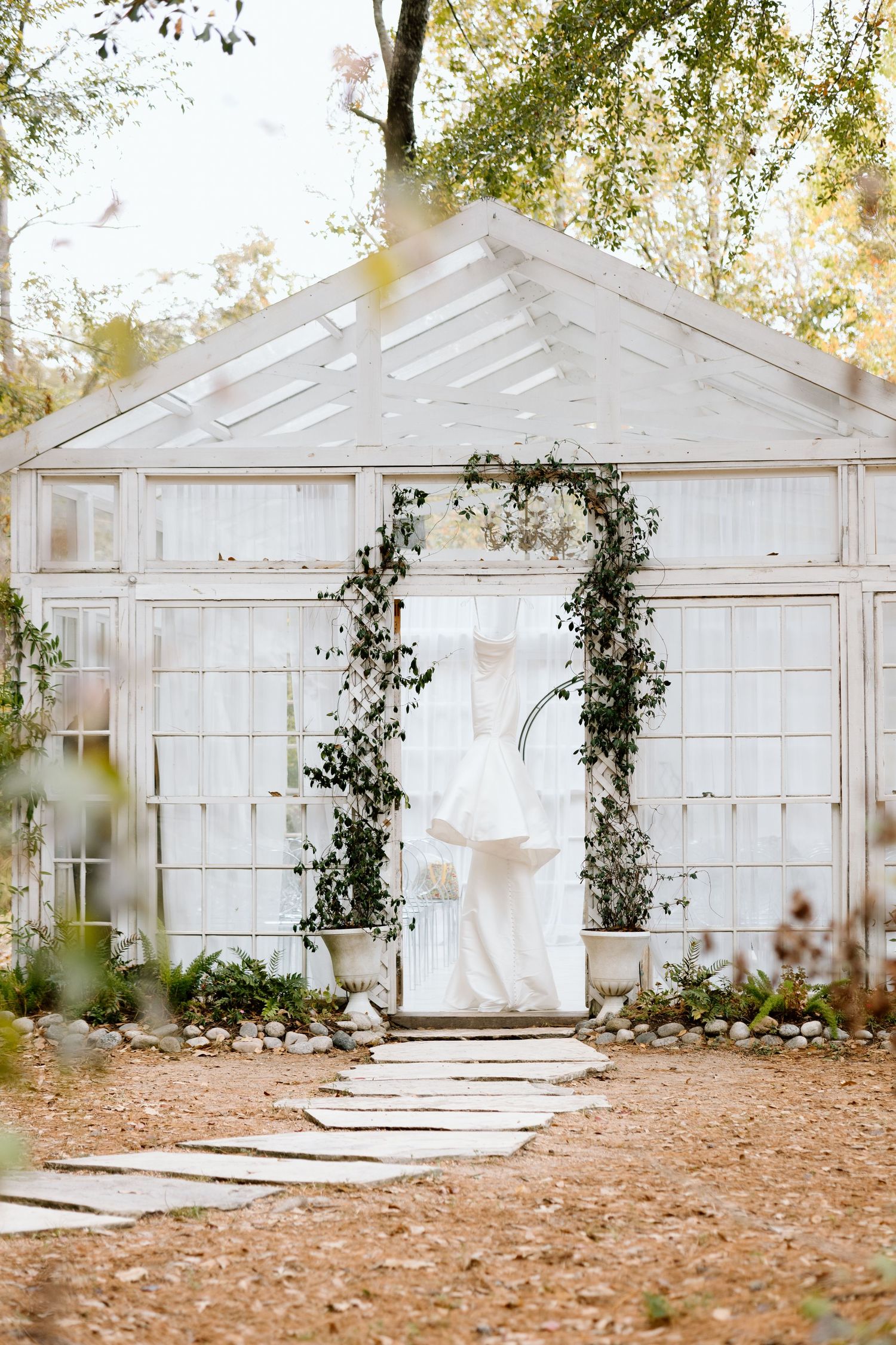 White greenhouse wedding venue adorned with floral arch and hanging bridal gown surrounded by autumn trees.