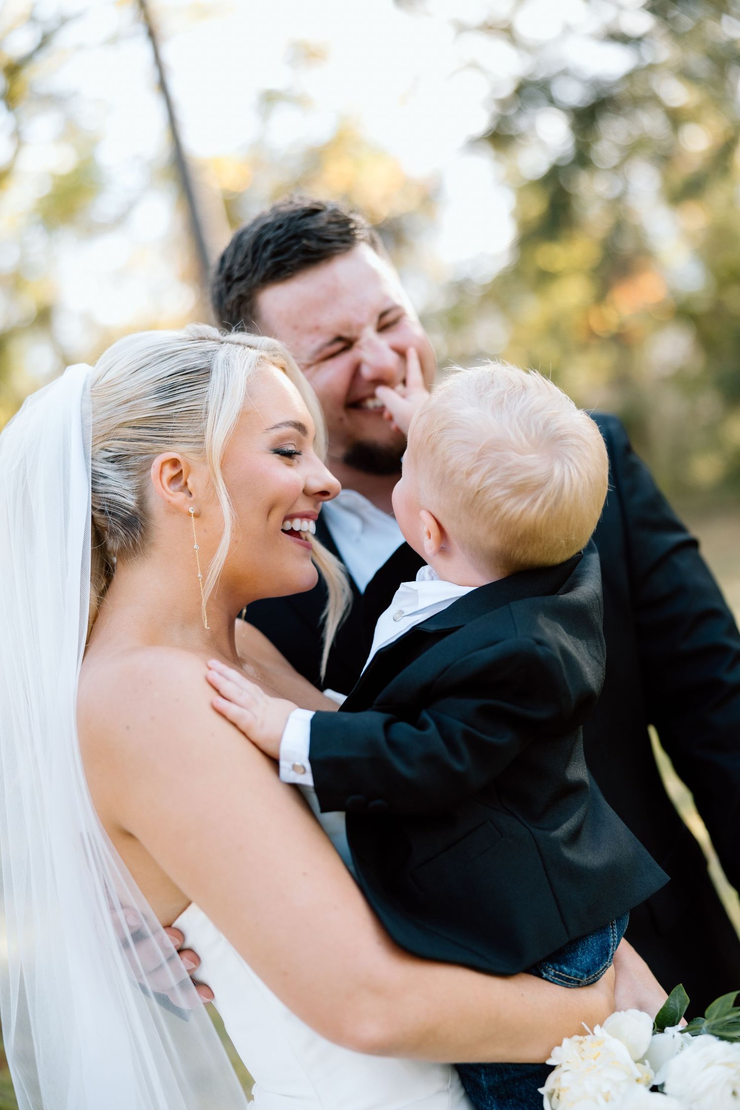 Bride and groom in wedding attire laughing with a young toddler boy in a black suit outdoors.