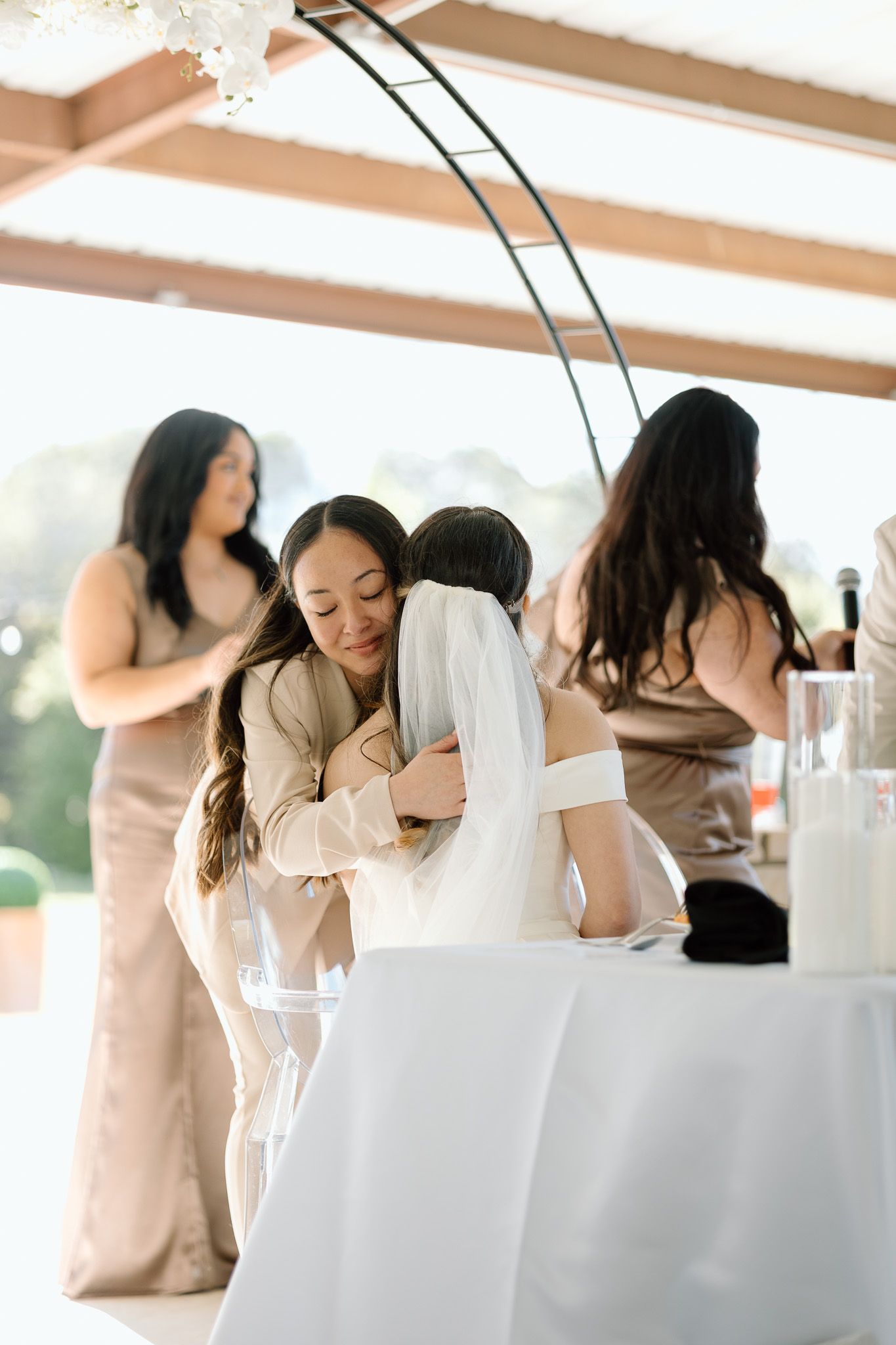 Wedding guests share emotional hugs and embraces during reception celebration under white tent.