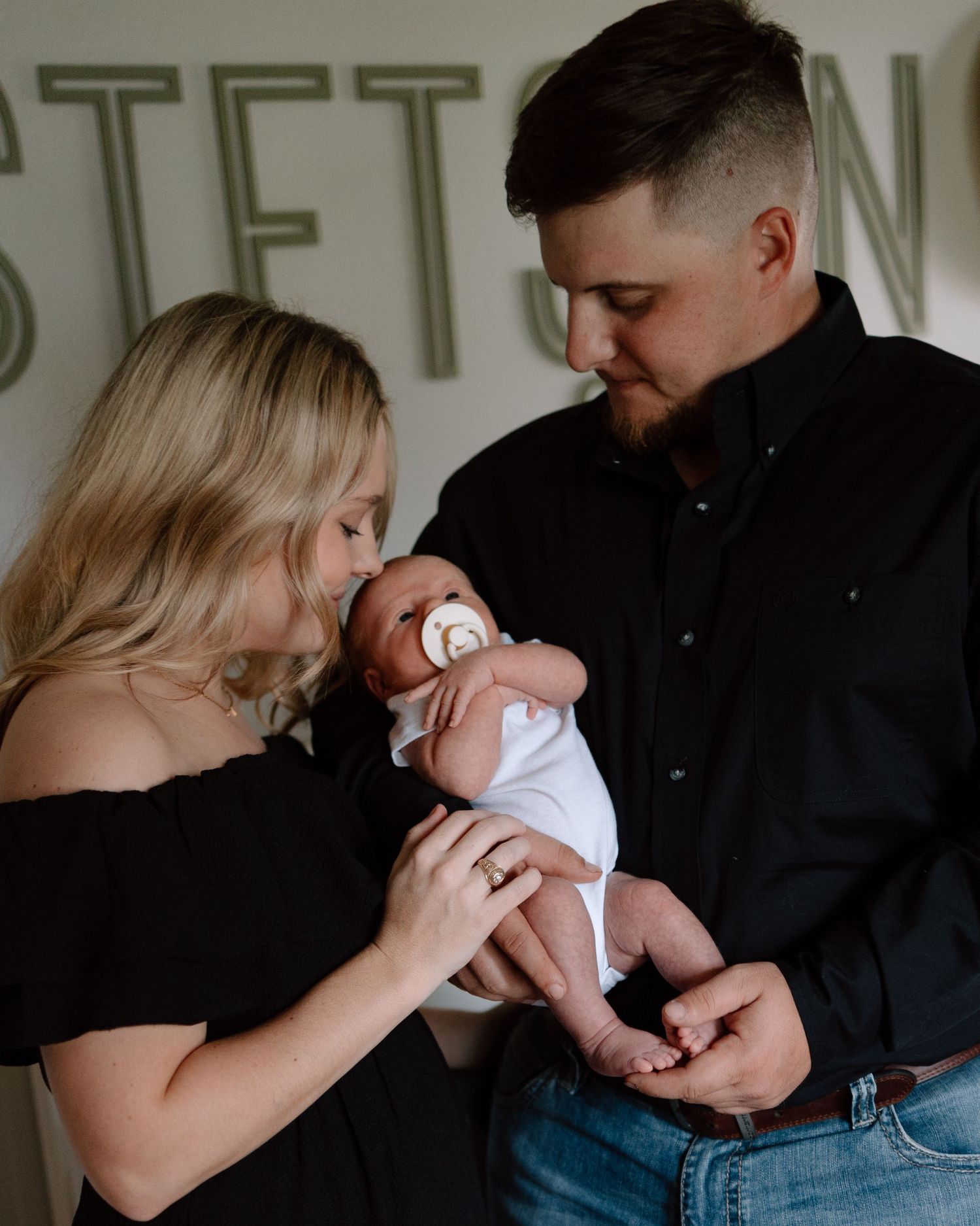 Two people in black outfits hold and gaze lovingly at a small baby in white clothing against a light wall with lettering.