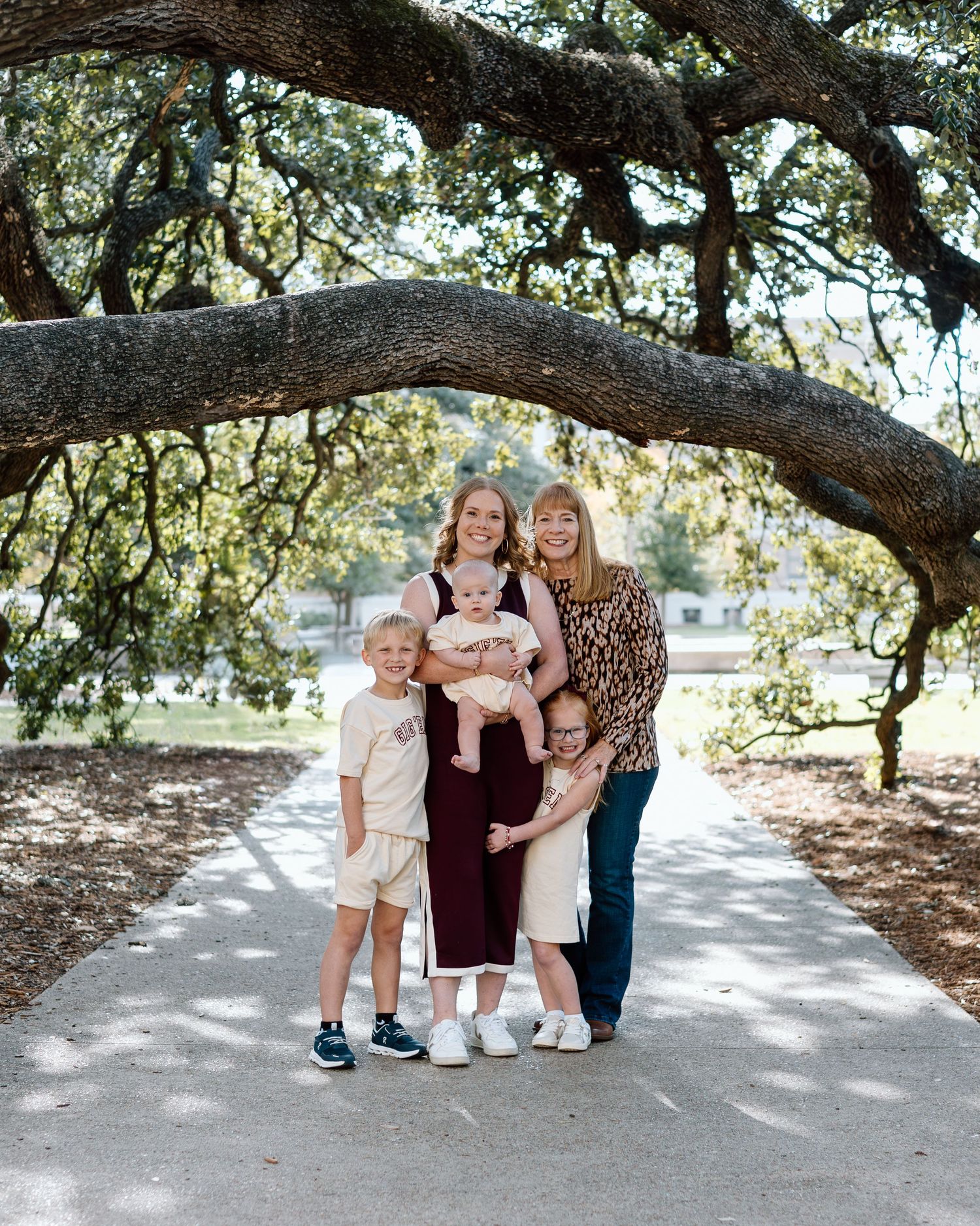 Family of four posing under a large oak tree on a sunny day, with two women holding children on a park path.