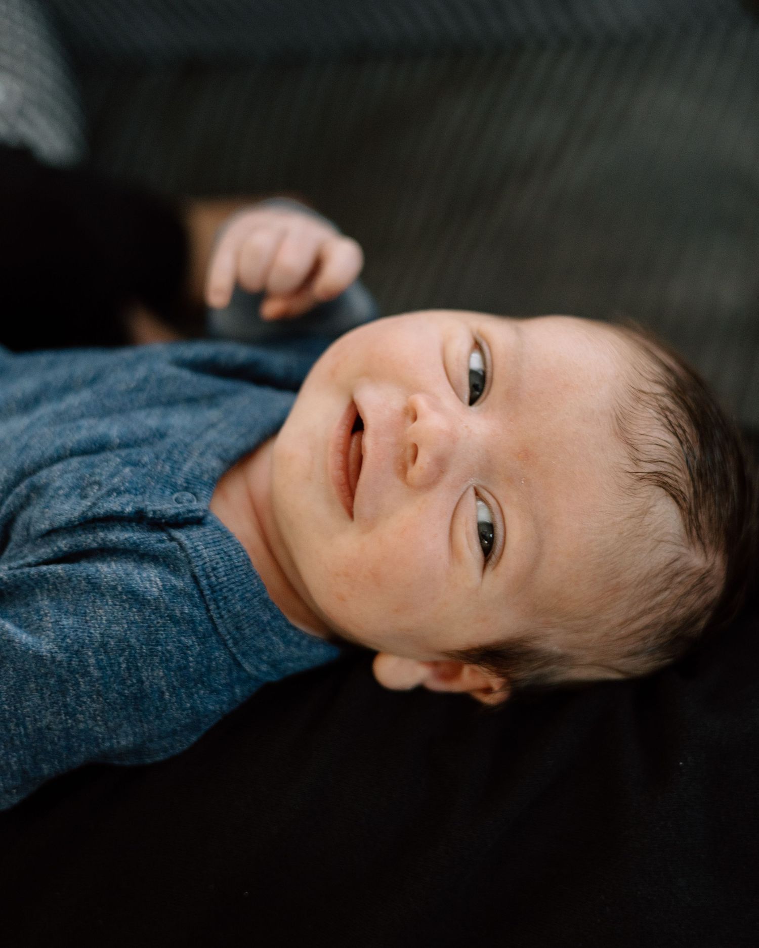 A smiling newborn baby with dark hair wearing a blue onesie, lying on a dark surface.