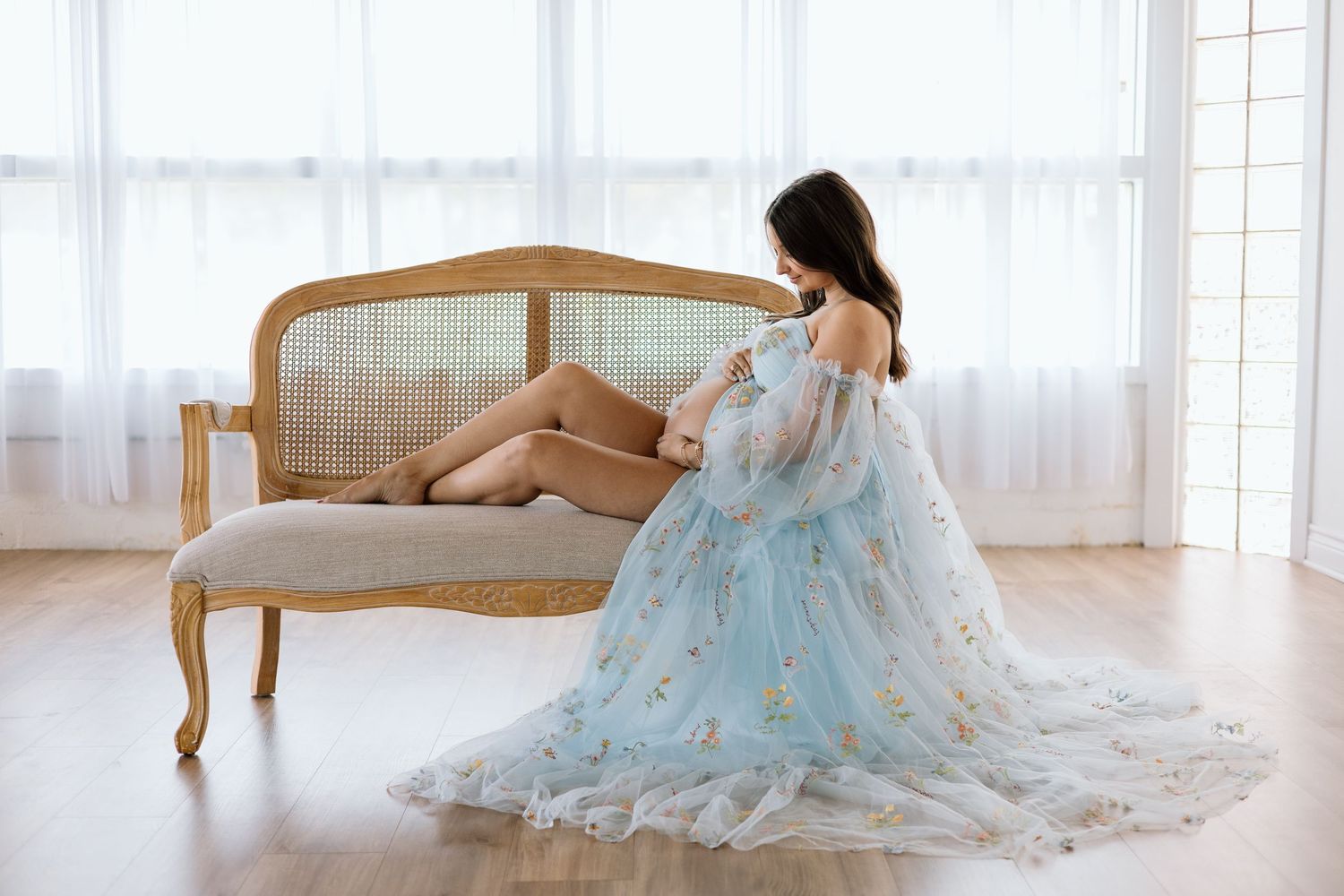 Pregnant woman in a flowing light blue floral gown sitting gracefully on a vintage sofa in a bright, airy room.