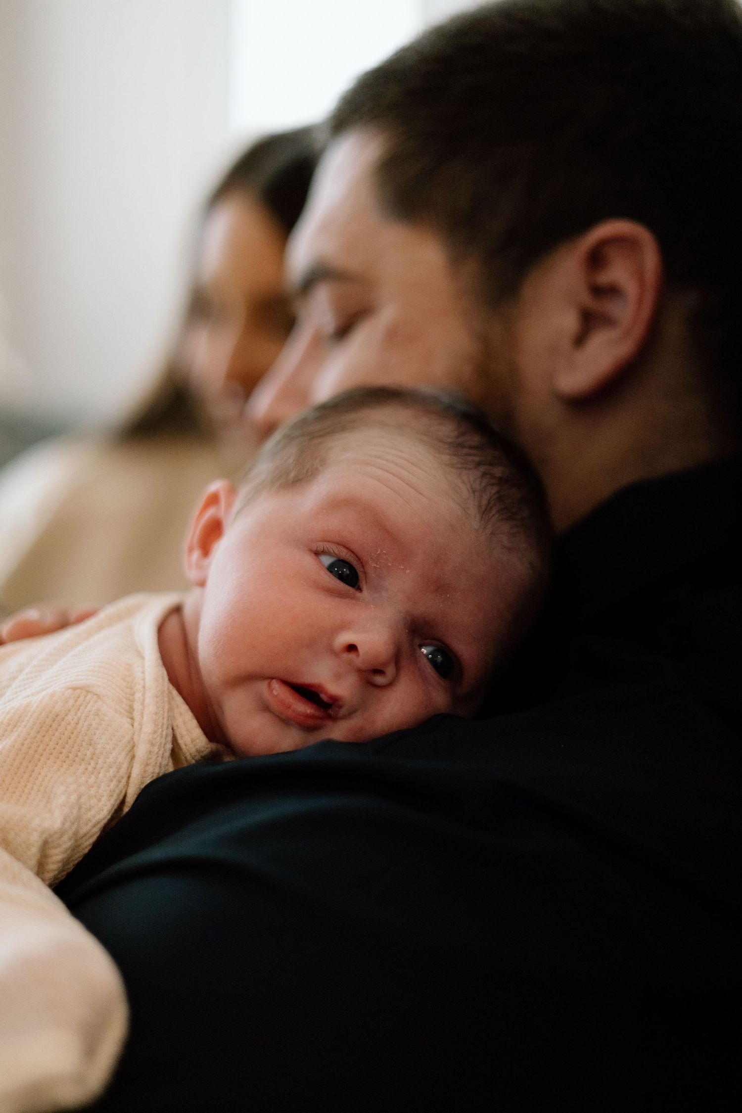 Close-up of newborn baby resting on fathers shoulder in dark shirt with mother visible in background.