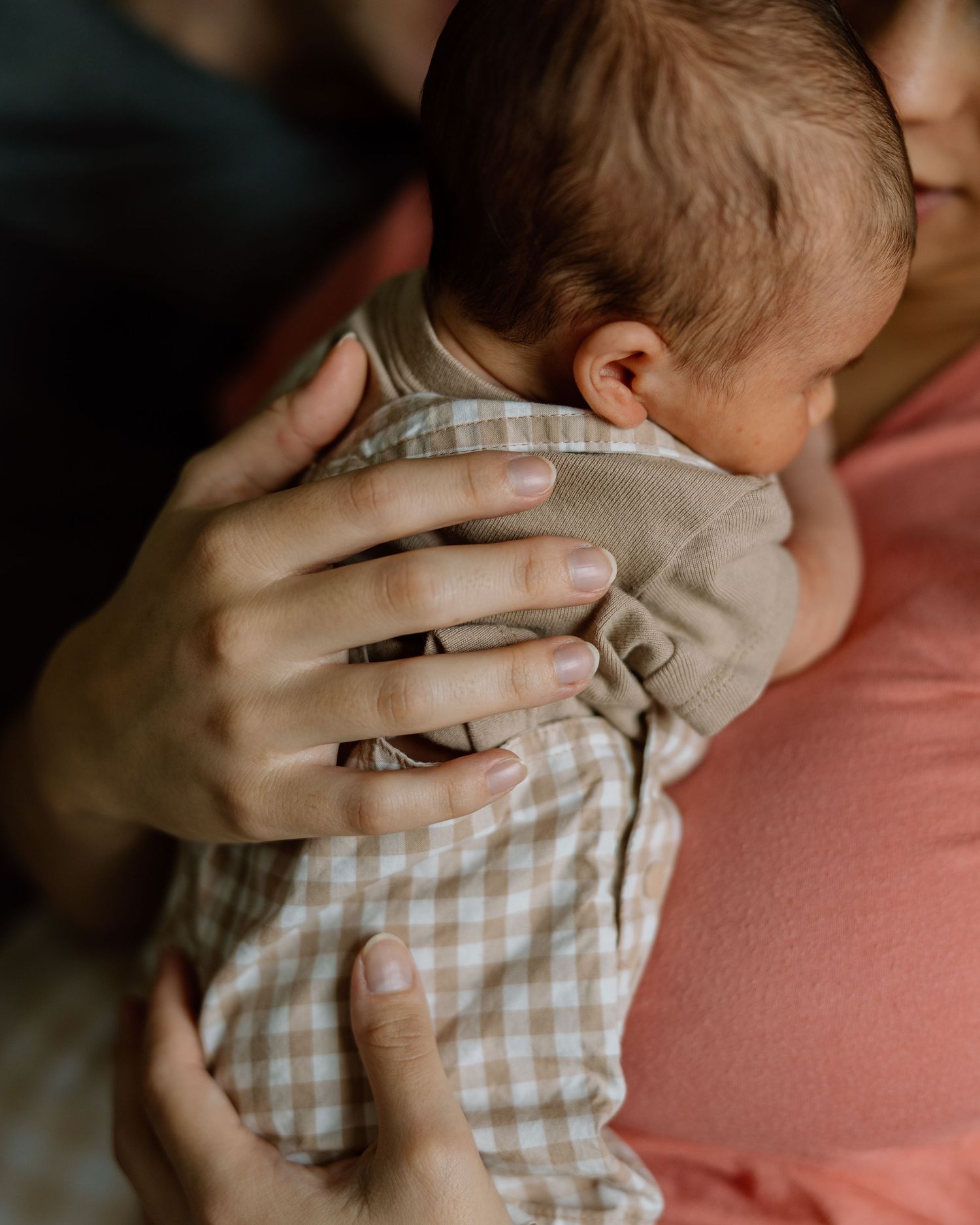 Close-up of parents' hands gently holding a newborn baby in a gingham outfit.
