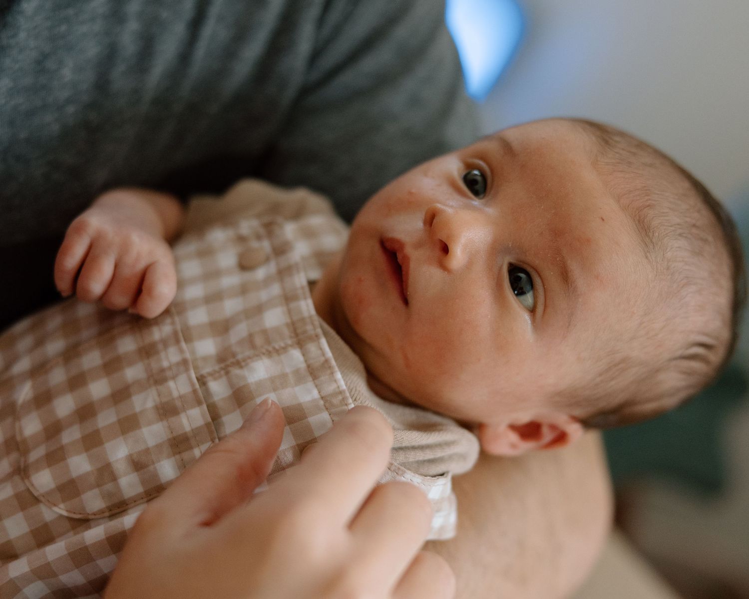 Newborn baby in gingham onesie being held by parent, looking up with curious eyes in a warm indoor setting.
