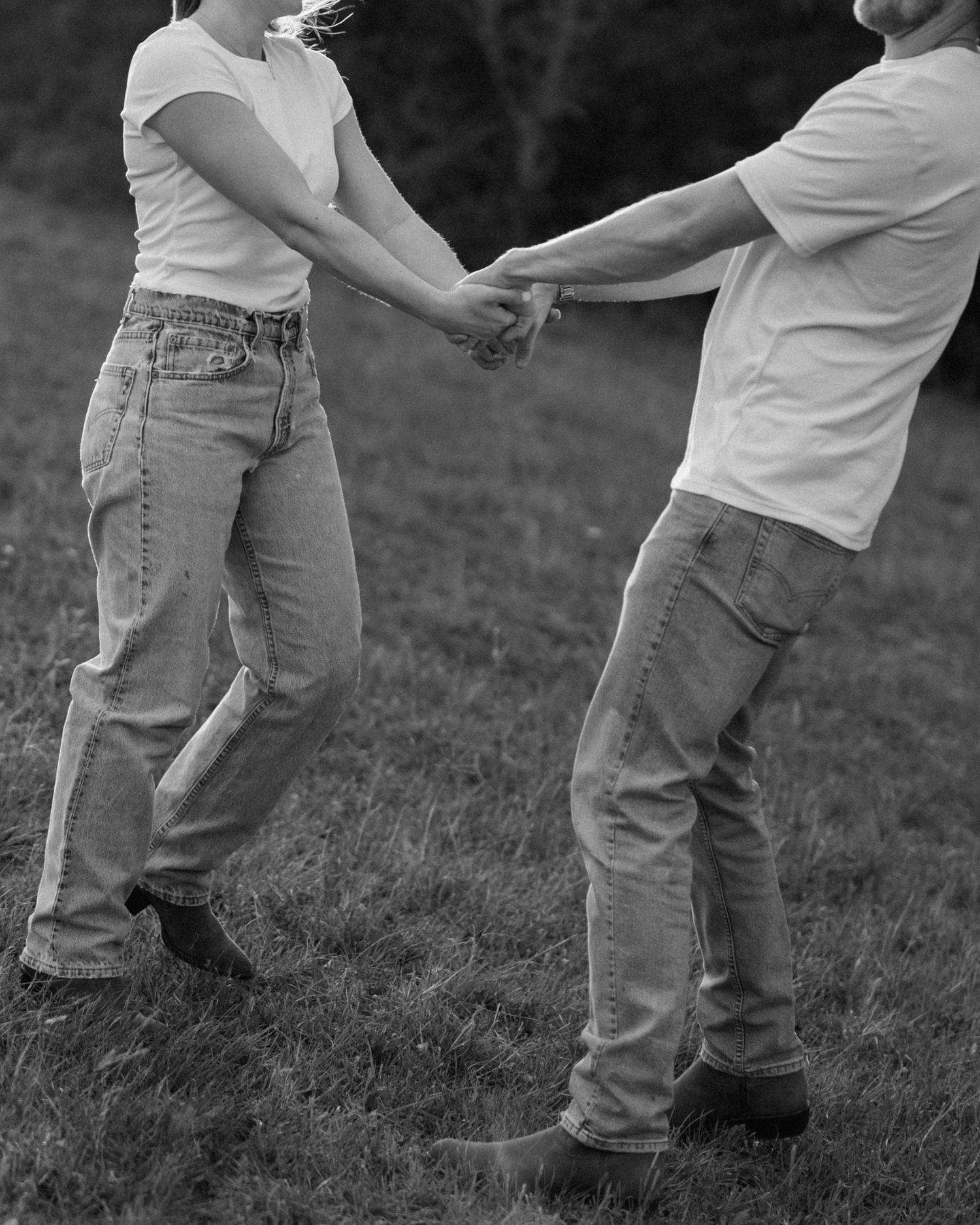 Black and white photo of couple playfully holding hands and dancing together on a grassy lawn.