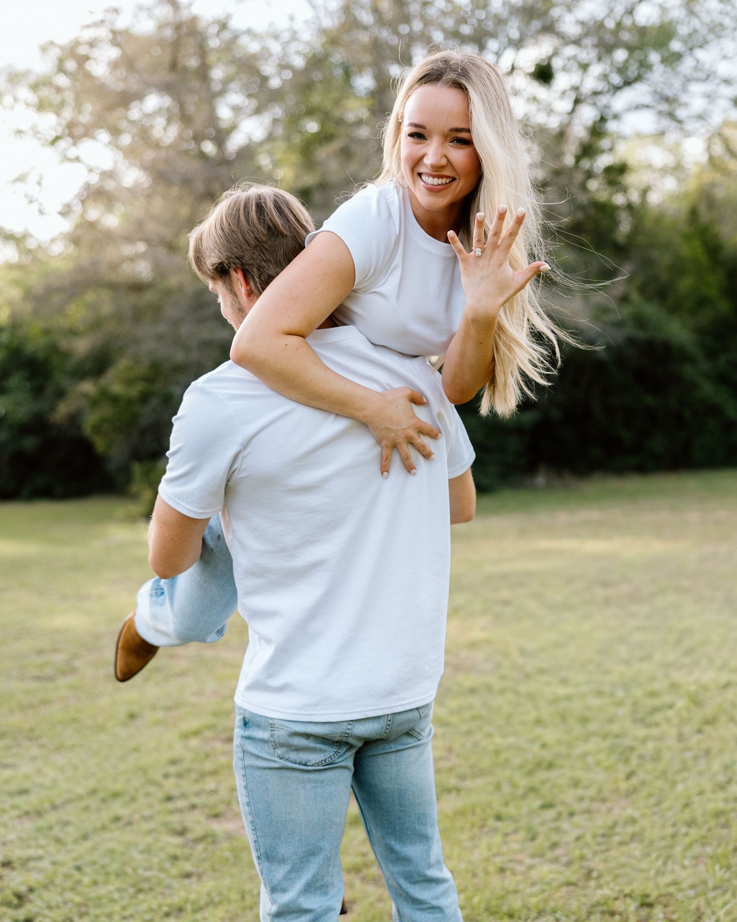 Joyful woman being lifted by her partner in a sunny park, showing off her engagement ring with a big smile.