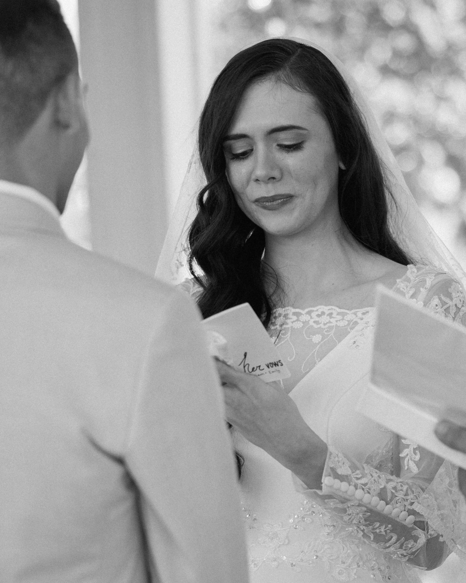 Black and white photo of an emotional bride reading wedding vows during her ceremony in a lace gown.