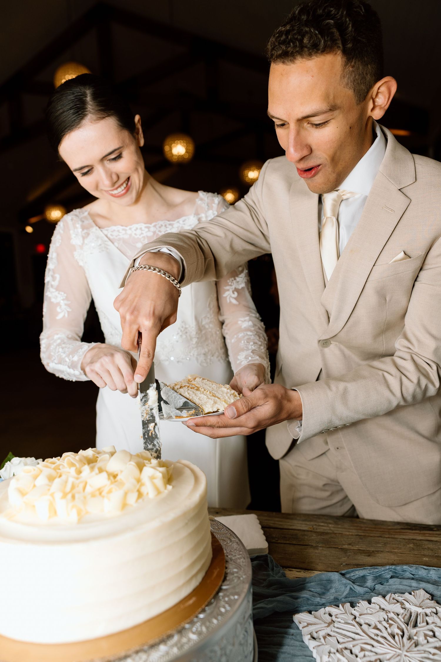 Bride and groom cut white wedding cake together at reception, groom in tan suit, bride in lace gown.