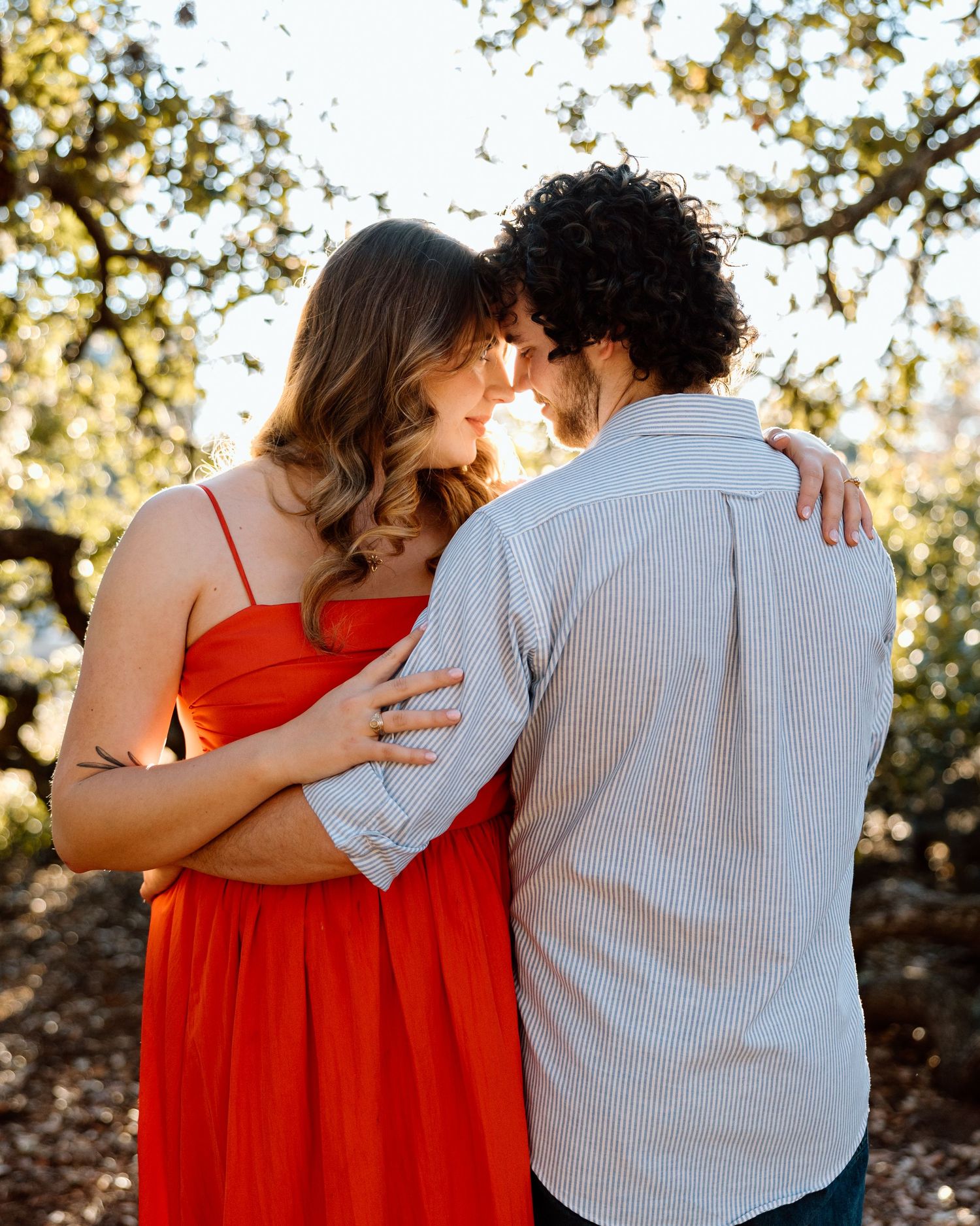 Couple in romantic pose outdoors, woman in red dress embracing man in striped shirt under sunlit trees.
