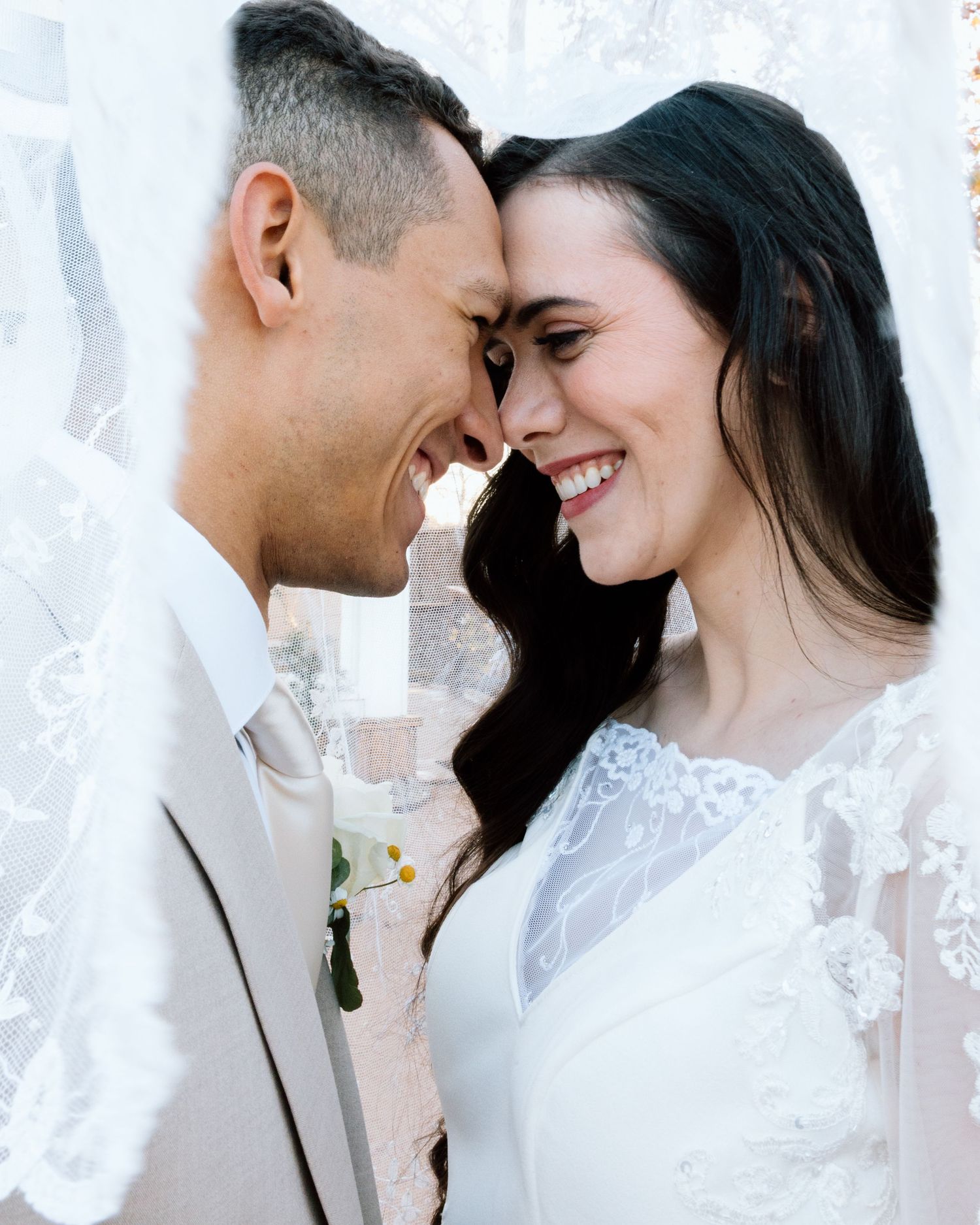 Smiling bride and groom touching foreheads under lace veil in romantic outdoor wedding portrait.