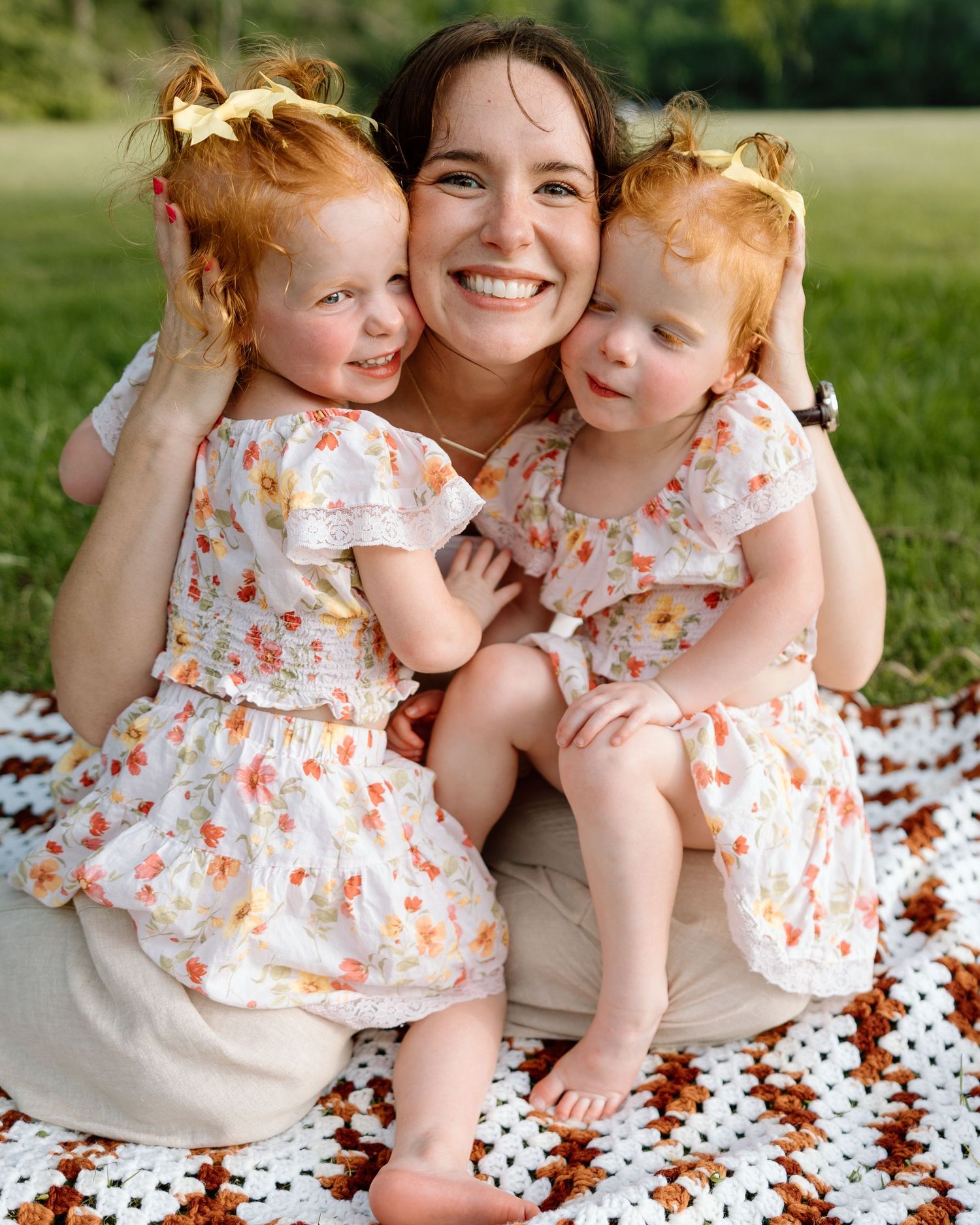 A smiling woman sits outdoors on a blanket hugging two young redheaded twin girls in matching floral dresses.