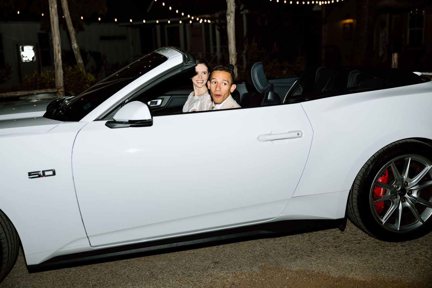 Newlyweds celebrating in white Ford Mustang 5.0 convertible after wedding reception.