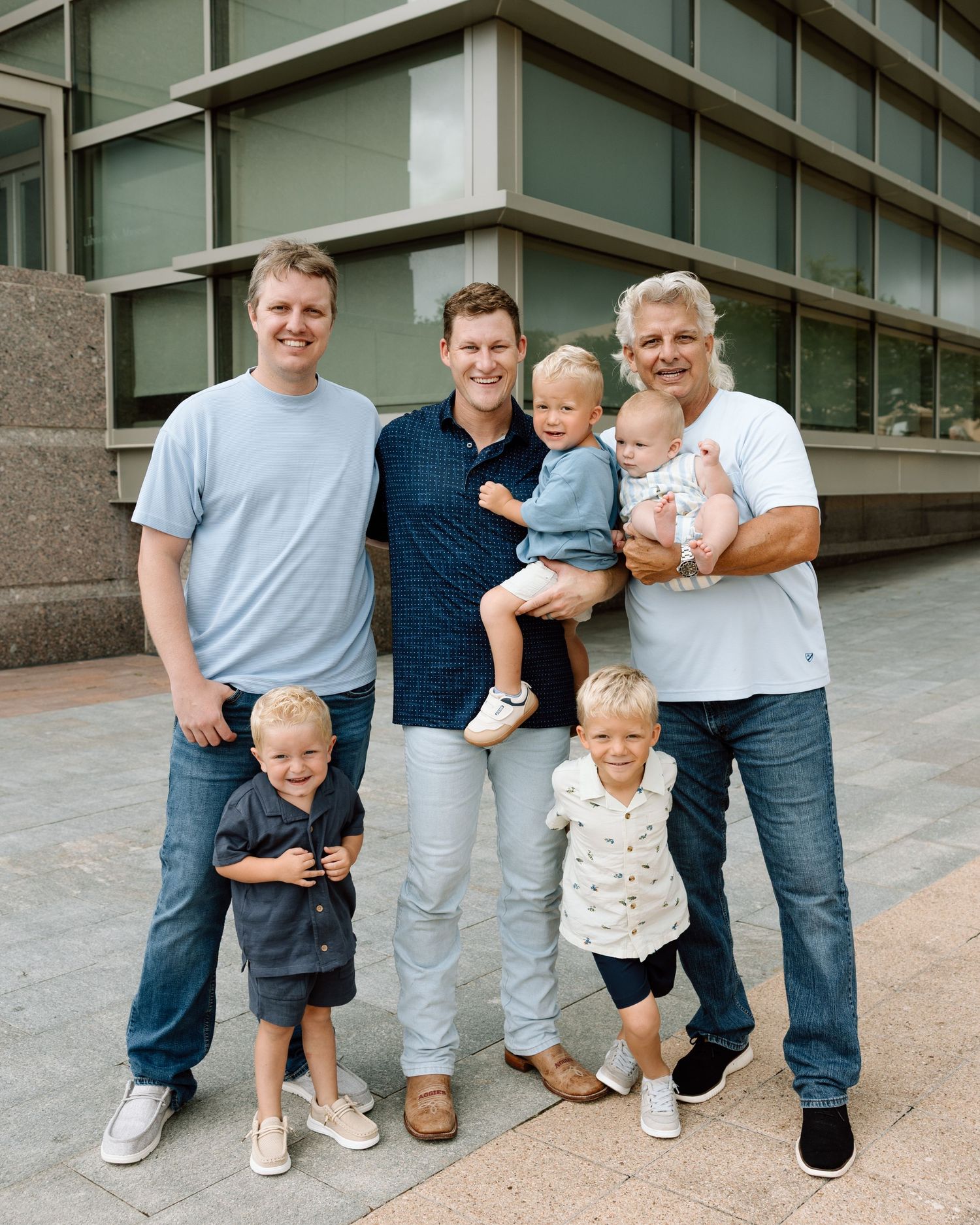 Three generations of men posing outdoors with young children and a baby in an urban setting.