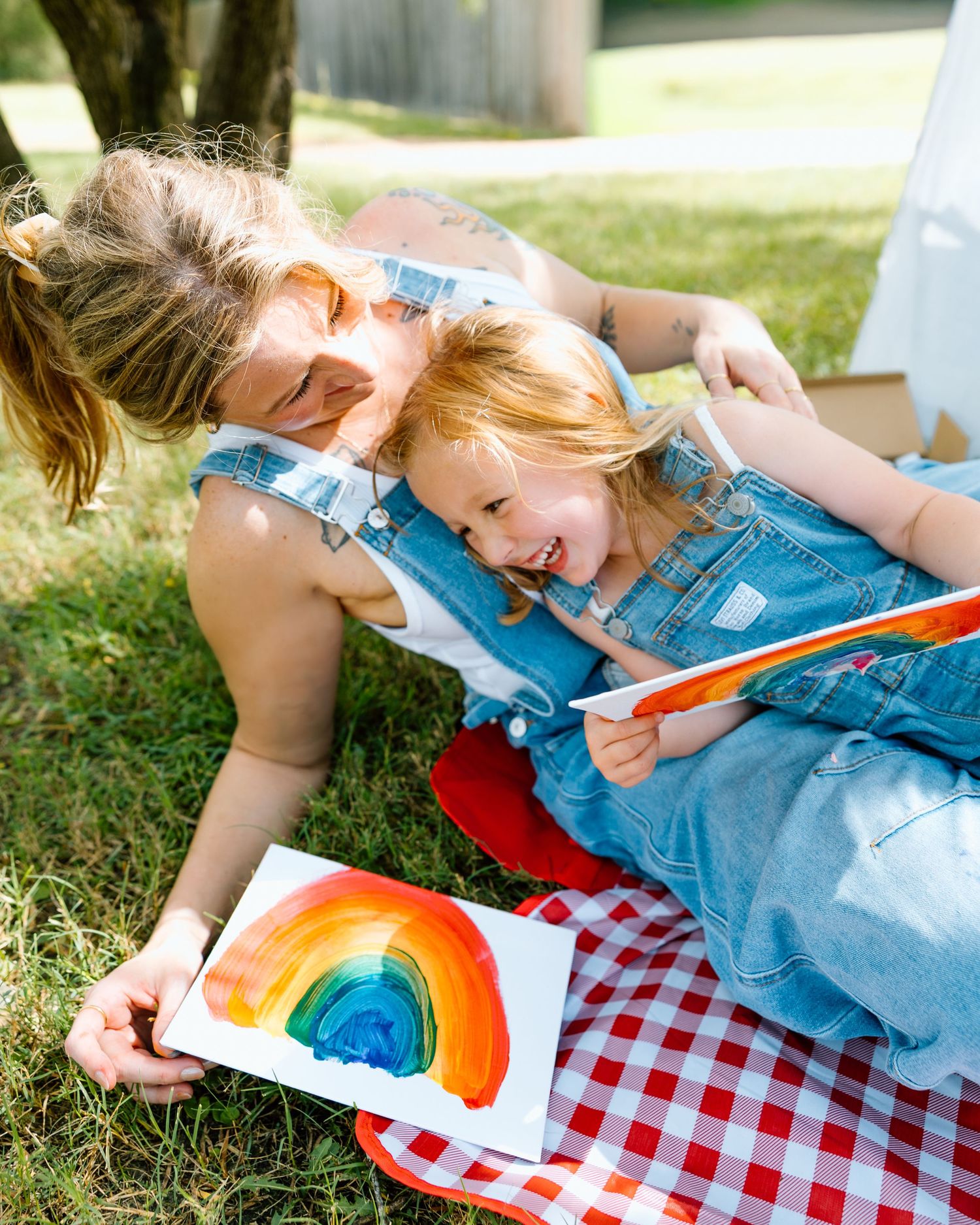 A mother and child laugh together on a picnic blanket outdoors, admiring a colorful rainbow painting.