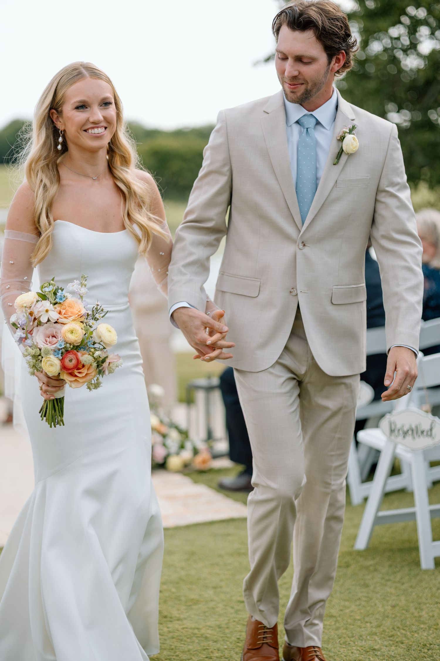 A couple walks hand in hand at their outdoor wedding, with the bride holding a peach and white bouquet.