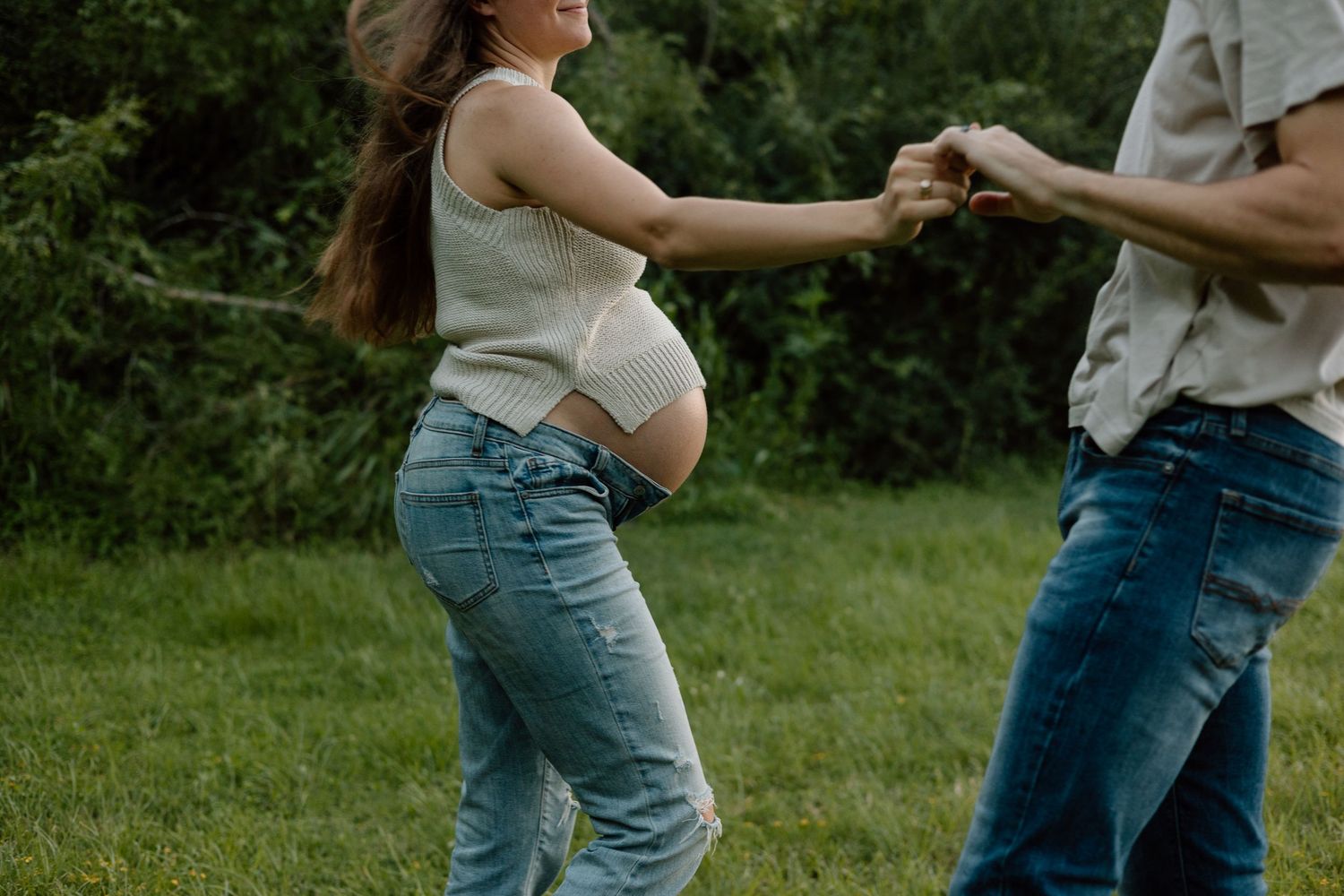 Dancing couple in casual wear celebrate their pregnancy in a green outdoor setting.