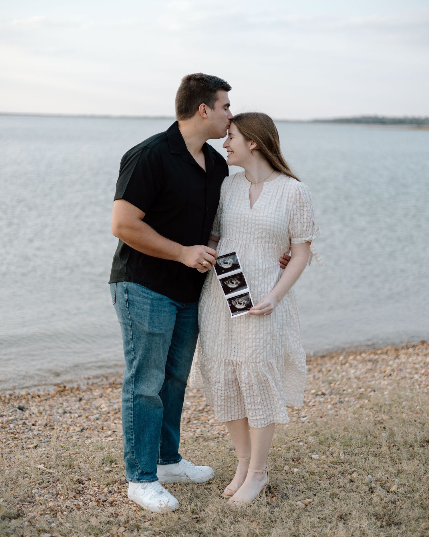 A couple poses together on a beach shoreline while displaying their pregnancy announcement photos.