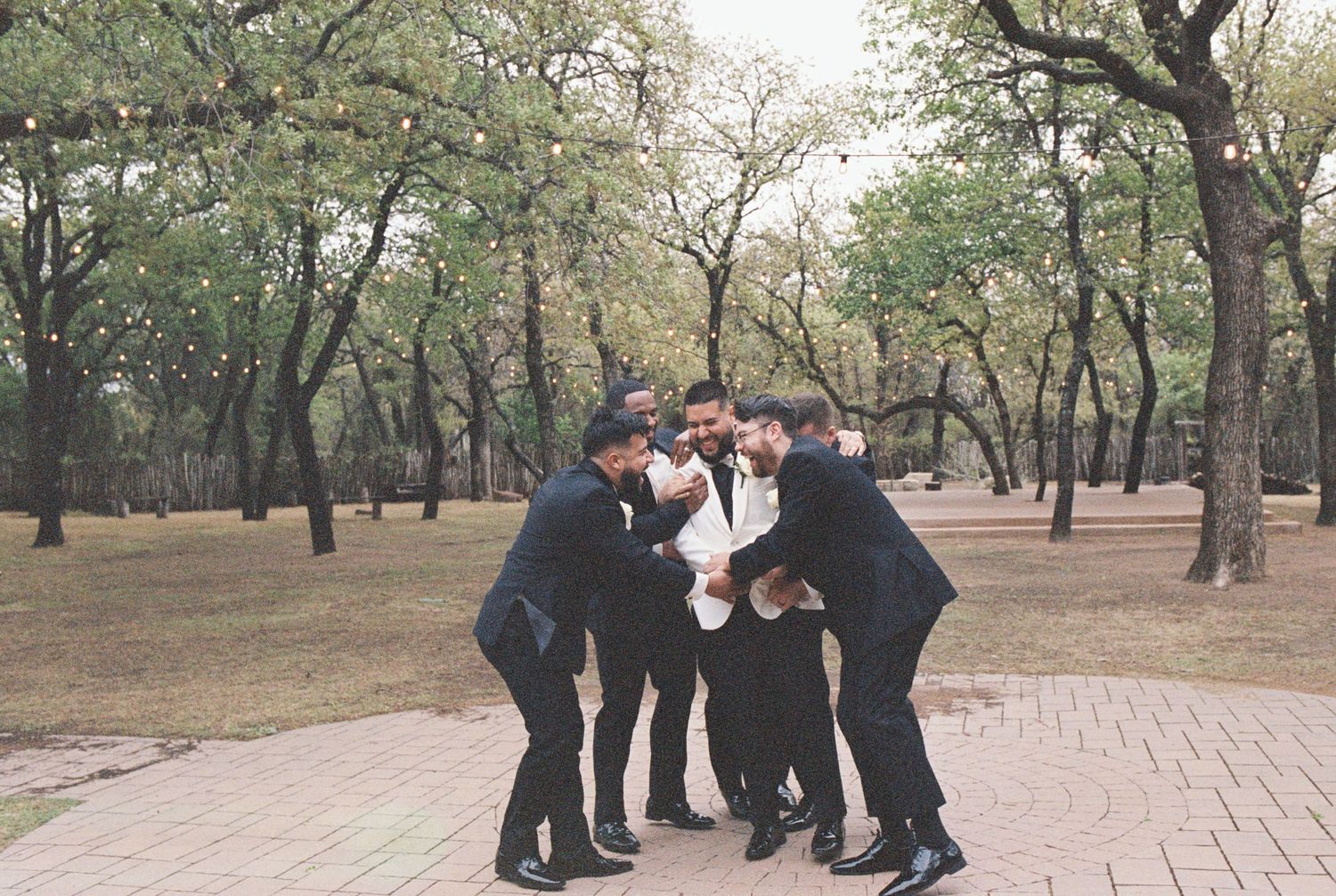 A group in formal attire huddles together playfully on a brick pathway in a wooded area.